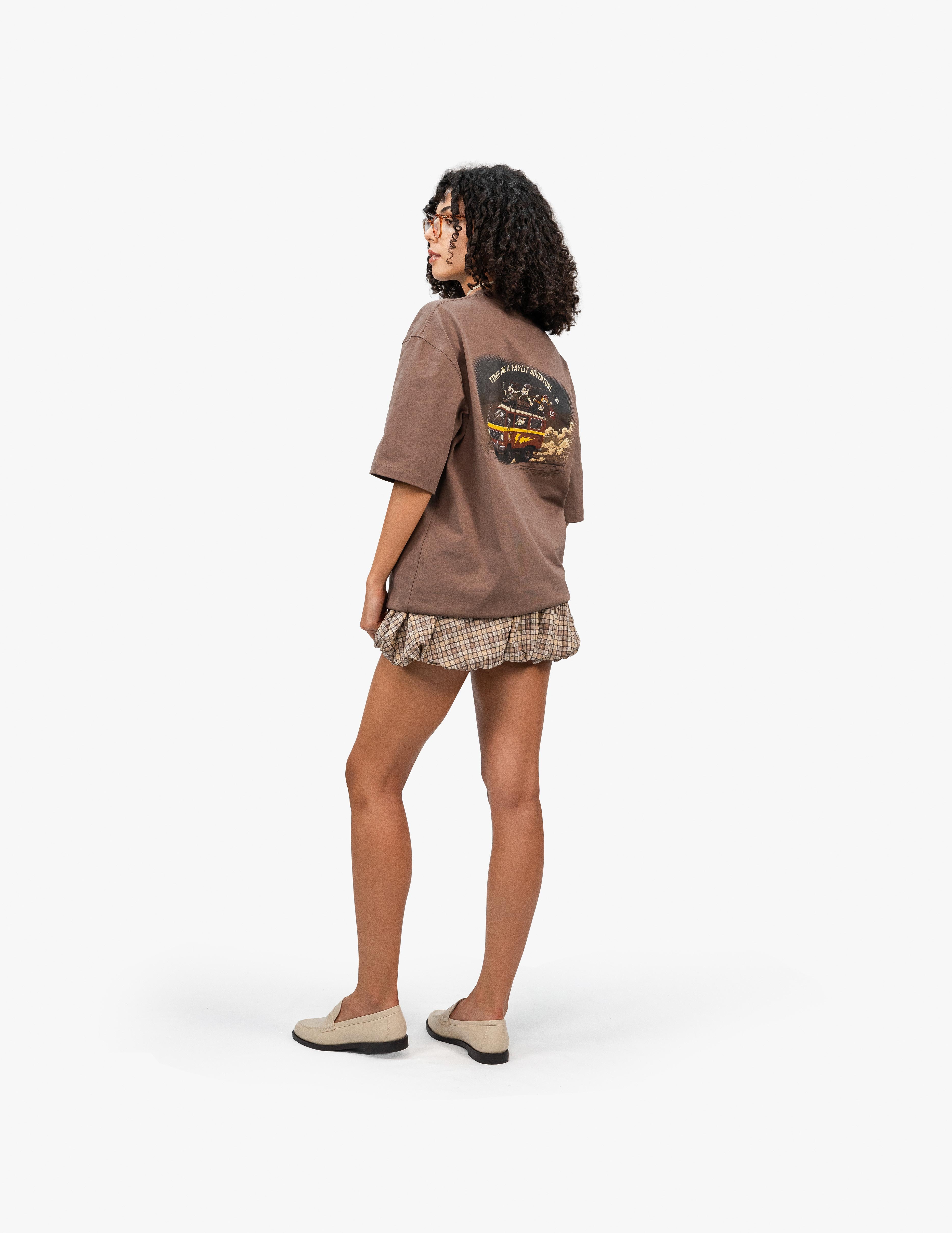 Adventure Kahverengi Oversize Unisex Tişört, Beyaz bir stüdyo fonunun önünde duran, kısa siyah kıvırcık saçlı ve gözlüklü bir kadın. Arkası dönük bir şekilde sol tarafa bakıyor. Üzerinde sırtında renkli bir grafik baskı bulunan kahverengi, geniş kesim bir tişört ve etek ucu fırfırlı, kahverengi ekose desenli bir şort giyiyor. Ayaklarında açık renk loafer ayakkabılar var. -  Faylit