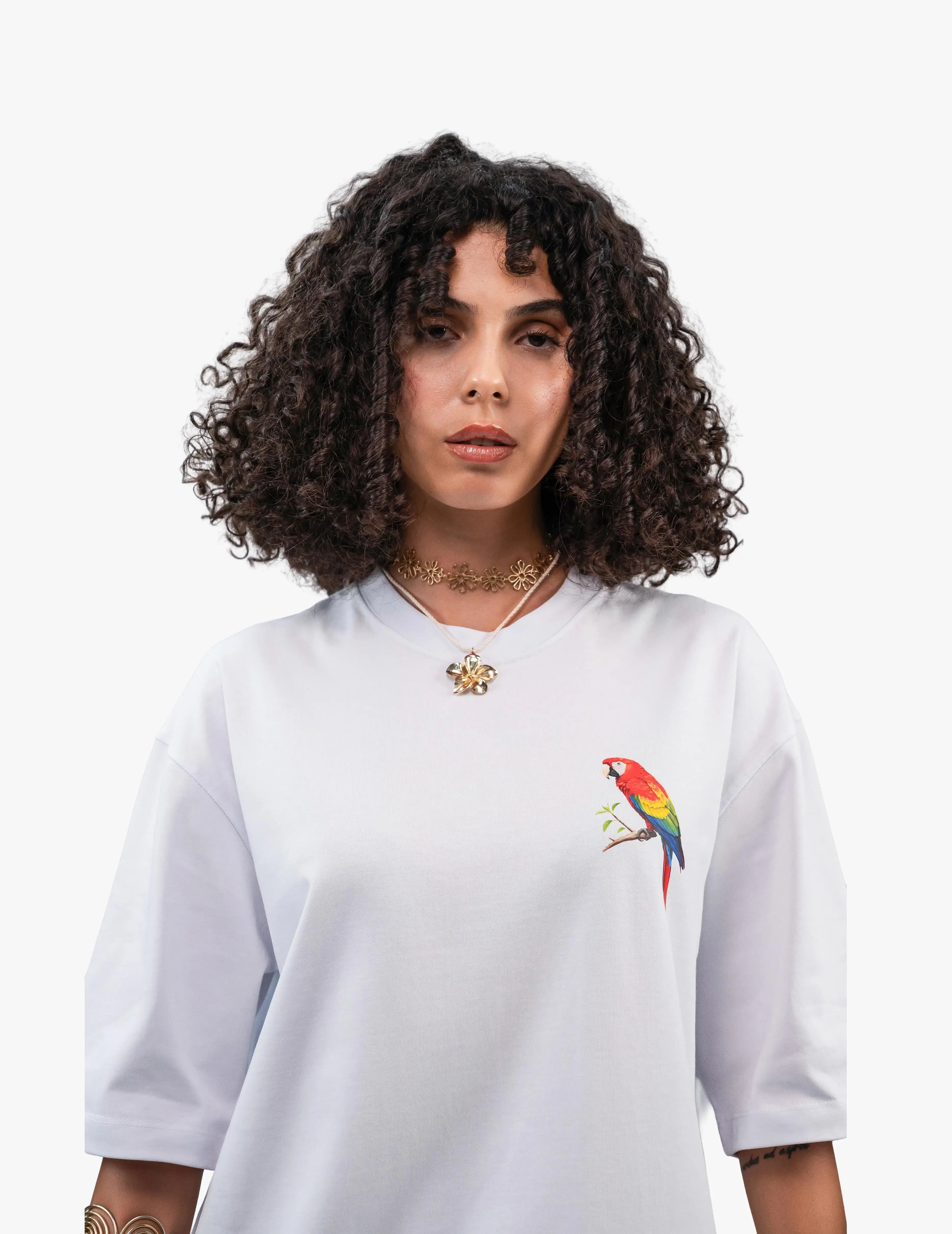 Beach Vibes Optik Beyaz Oversize Unisex Tişört, Beyaz bir fonun önünde, doğrudan kameraya bakan, kısa siyah kıvırcık saçlı bir kadın. Üzerinde sağ göğüs hizasında renkli bir papağan figürü bulunan beyaz, geniş kesim bir tişört giyiyor. Boynunda altın renkli, çiçek desenli bir gerdanlık ve beyaz boncuklu bir kolye takıyor. -  Faylit