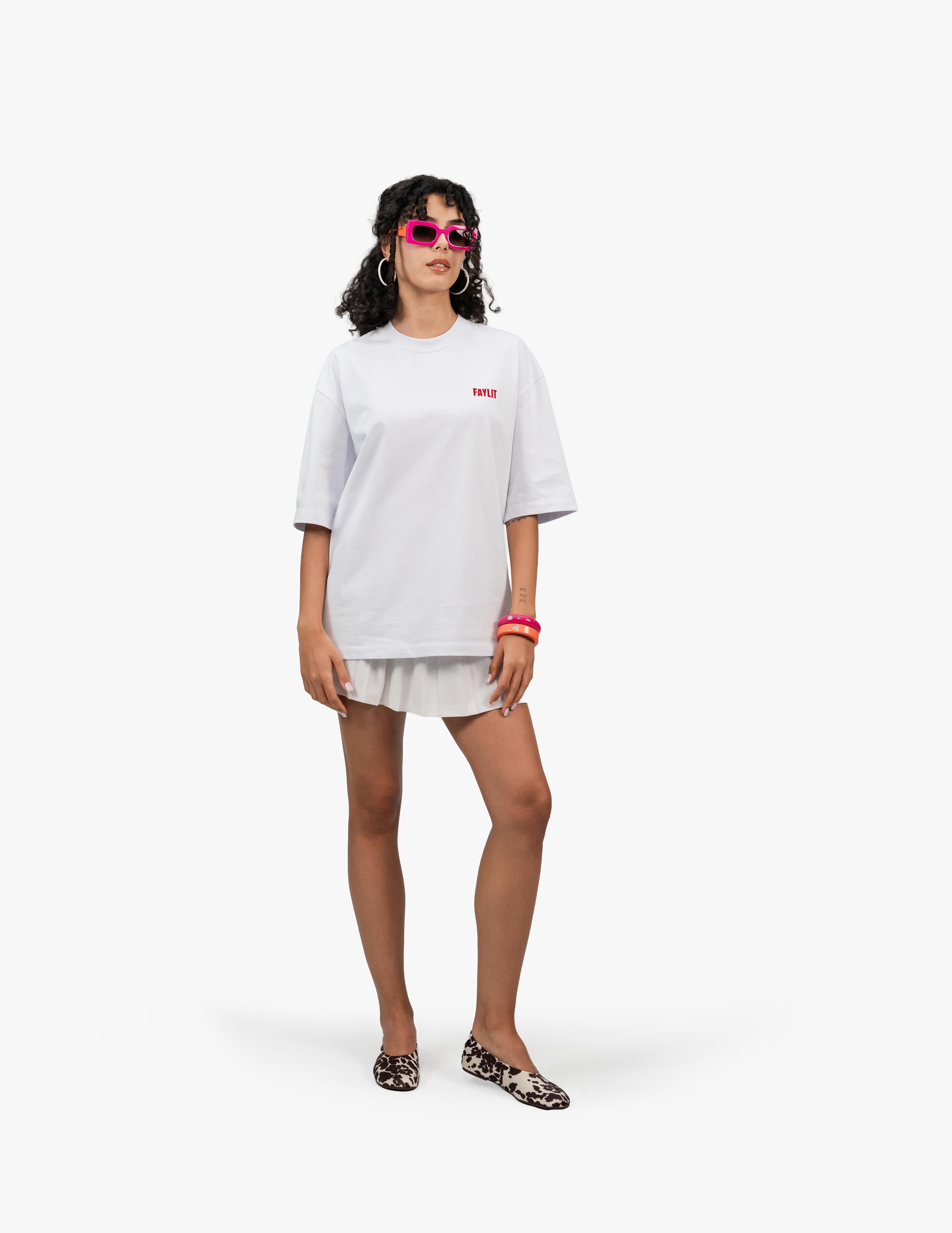 FDNA_03 Oversize Tişört,  Beyaz bir stüdyo fonunun önünde, hafifçe sağa dönük duran, kısa siyah kıvırcık saçlı bir kadın. Pembe çerçeveli güneş gözlüğü ve büyük gümüş renkli küpeler takıyor. Üzerinde göğüs kısmında küçük pembe bir yazı bulunan beyaz, geniş kesim bir tişört ve beyaz pileli bir mini etek giyiyor. Sol kolunda pembe ve turuncu bileklikler var. Elleri yanlarda, hafifçe aşağıda duruyor. Ayaklarında leopar desenli babet ayakkabılar bulunuyor. -  Faylit