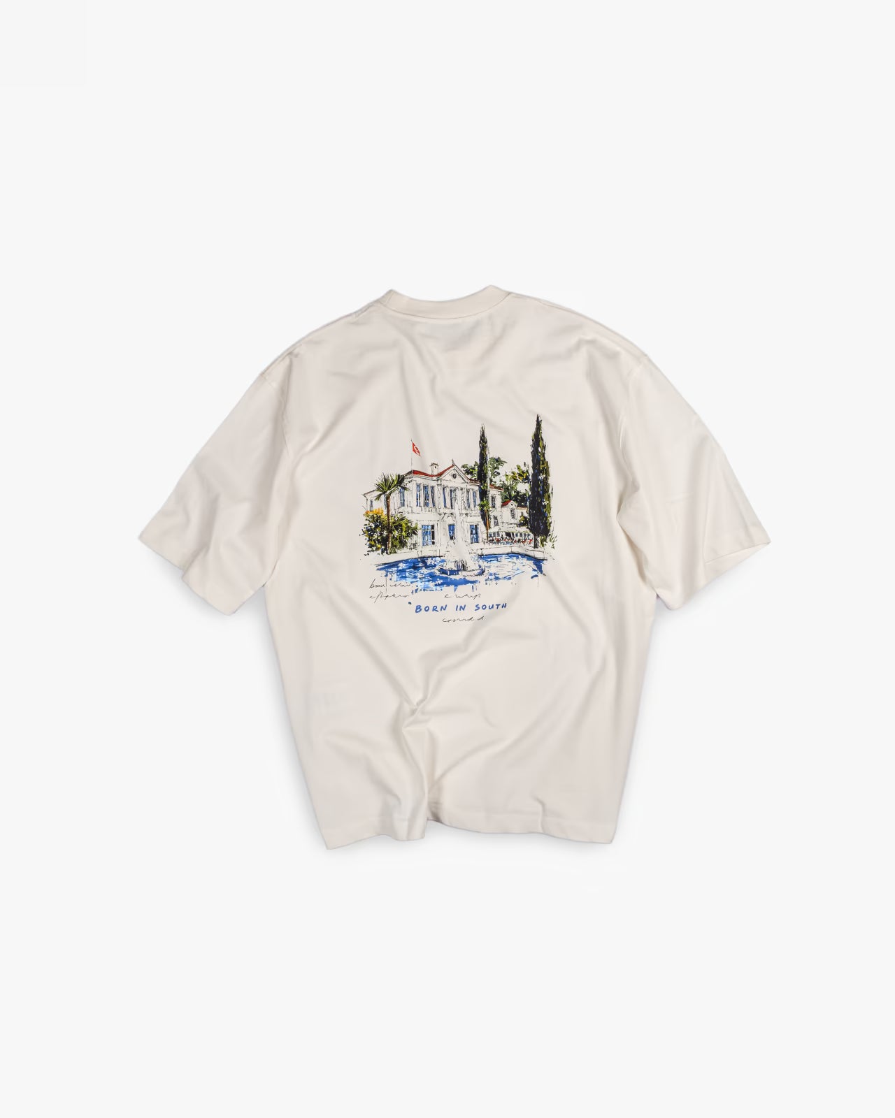 FAYLIT kırık beyaz oversize tişört, arka kısmında Born in South yazılı, havuzlu ve ağaçlı, Akdeniz stili ev içeren renkli grafik baskı, unisex ve boxy fit kesim.