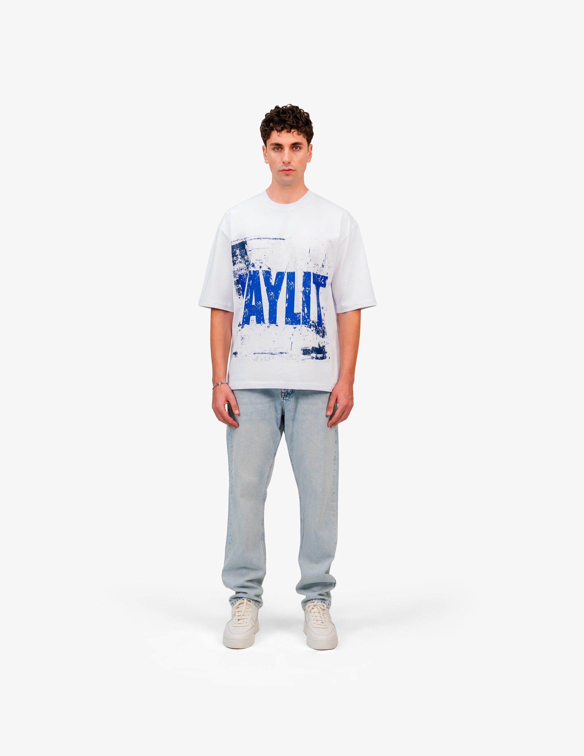 00Blue Optik Beyaz Oversize Tişört Beyaz stüdyo fonunda, tam karşıdan poz veren erkek manken. Üzerinde, göğsünde mavi renkte, büyük ve eskitme (distressed) tarzda "FAYLIT" yazılı beyaz oversize bir tişört, açık renkli, bol kesim bir kot pantolon ve beyaz spor ayakkabılar var. -  Faylit