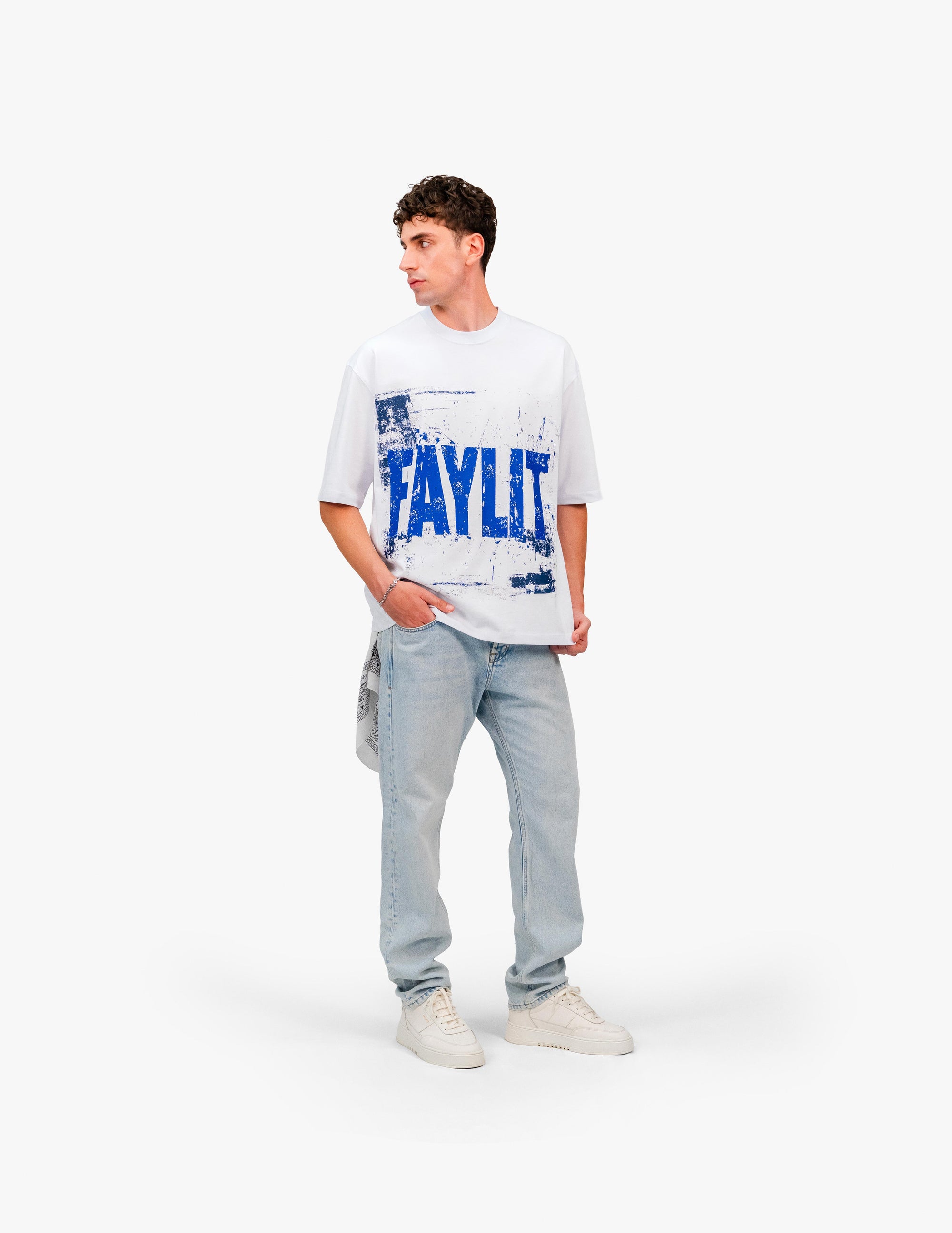00Blue Optik Beyaz Oversize Tişört Beyaz stüdyo fonunda, yana dönük poz veren erkek manken. Üzerinde, mavi "FAYLIT" baskılı beyaz oversize tişört ve açık renkli, bol kesim kot pantolon giyiyor. Kombin, beyaz spor ayakkabılarla tamamlanmış. Arka cebinden desenli bir mendil/bandana sarkıyor. -  Faylit