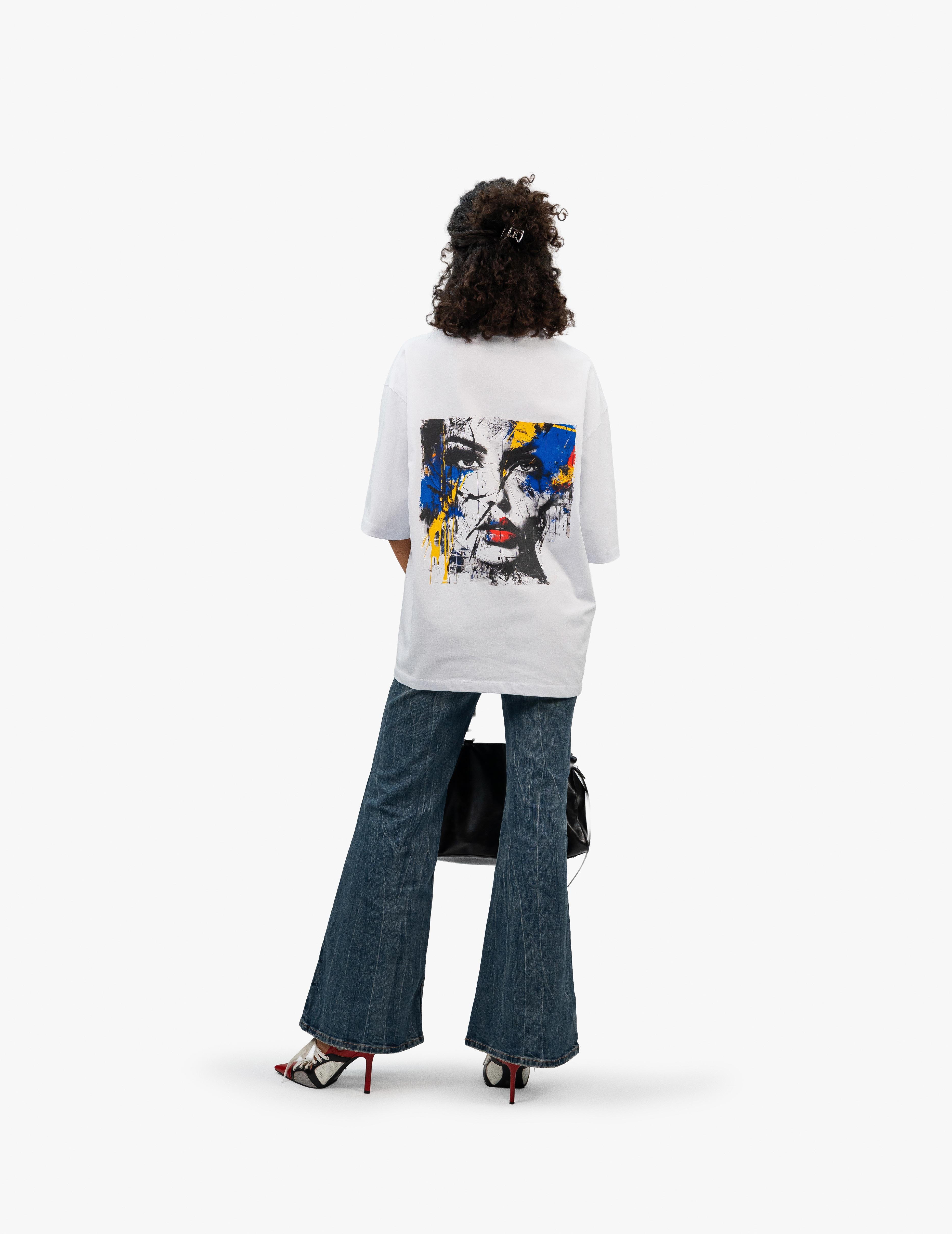 Abstract Portrait Grafik Beyaz Oversize Tişört, Beyaz bir stüdyo fonunun önünde, arkası dönük ve hafifçe sola dönük bir şekilde duran, kısa siyah kıvırcık saçlı bir kadın. Üzerinde sırt kısmında siyah beyaz bir kadın yüzü ve etrafında sarı, mavi, kırmızı renkli boya sıçramaları bulunan beyaz, geniş kesim bir tişört giyiyor. Altında bol paça, yıpranmış görünümlü bir kot pantolon ve kırmızı-siyah detaylı, topuklu ayakkabılar var. Sağ elinde siyah deri bir çanta tutuyor. -  Faylit