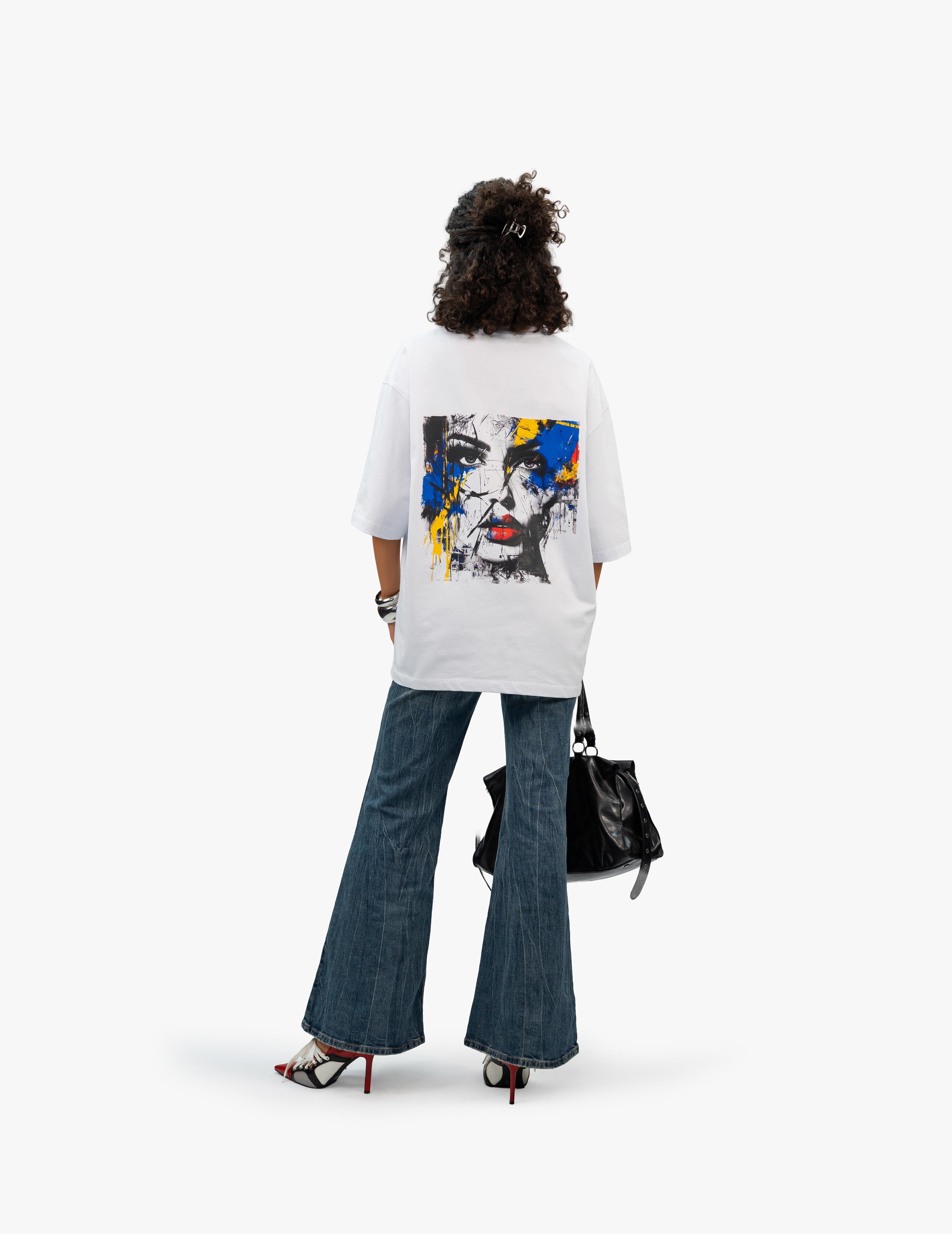 Abstract Portrait Grafik Beyaz Oversize Tişört, Beyaz bir stüdyo fonunun önünde, arkası dönük duran, kısa siyah kıvırcık saçlı bir kadın. Üzerinde sırt kısmında siyah beyaz bir kadın yüzü ve etrafında sarı, mavi, kırmızı renkli boya sıçramaları bulunan beyaz, geniş kesim bir tişört giyiyor. Altında bol paça, yıpranmış görünümlü bir kot pantolon ve kırmızı-siyah detaylı, topuklu ayakkabılar var. Sağ elinde siyah deri bir çanta tutuyor. -  Faylit
