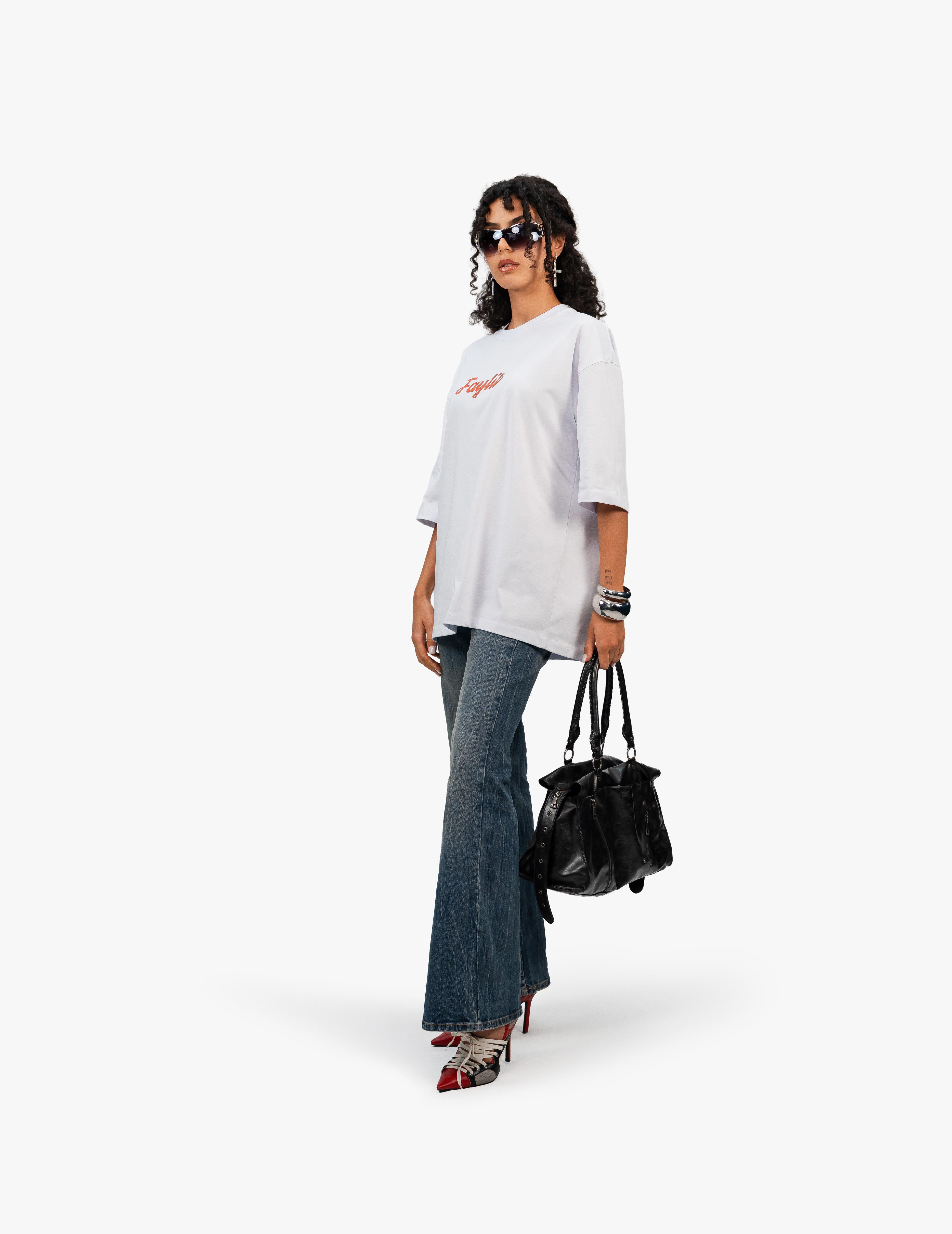 Abstract Portrait Grafik Beyaz Oversize Tişört, Beyaz bir stüdyo fonunun önünde, hafifçe sağa dönük ve aşağıdan çekilmiş bir açıyla duran, kısa siyah kıvırcık saçlı bir kadın. Siyah çerçeveli, dikdörtgen formda bir güneş gözlüğü takıyor. Üzerinde "Faylit" yazısı bulunan beyaz, geniş kesim bir tişört, bol paça, yıpranmış görünümlü bir kot pantolon ve kırmızı-siyah detaylı, topuklu ayakkabılar giyiyor. Sol kolunda bilezikler ve sağ elinde siyah deri bir çanta tutuyor. -  Faylit