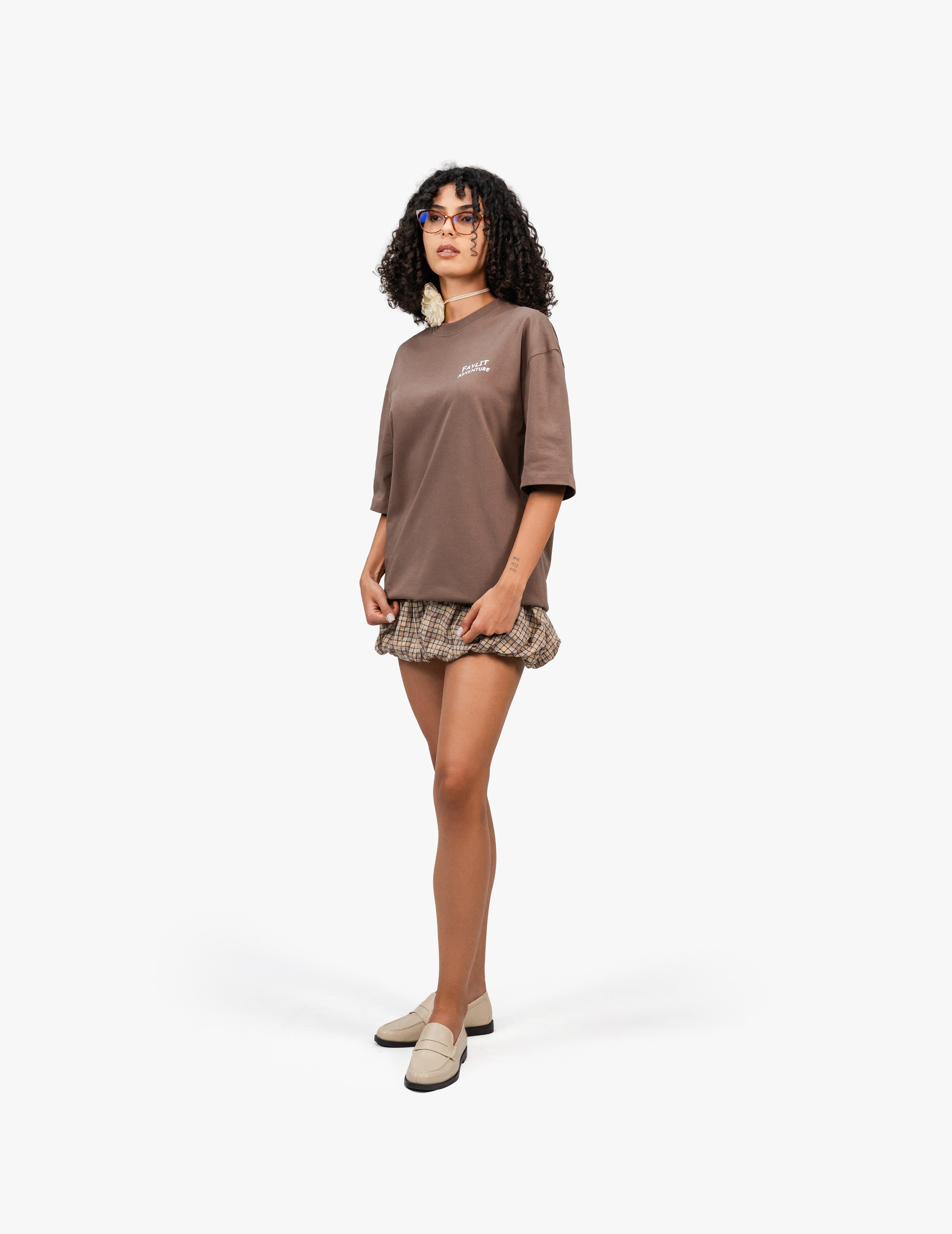 Adventure Kahverengi Oversize Unisex Tişört, Beyaz bir stüdyo fonunun önünde, hafifçe sağa dönük bir şekilde duran, kısa siyah kıvırcık saçlı ve gözlüklü bir kadın. Üzerinde sol göğüs hizasında beyaz bir yazı bulunan kahverengi, geniş kesim bir tişört ve etek ucu fırfırlı, kahverengi ekose desenli bir şort giyiyor. Sol eli cebinde, sağ eli ise yanında hafifçe aşağıda duruyor. Ayaklarında açık renk loafer ayakkabılar var ve boynunda beyaz bir çiçek detayı dikkat çekiyor. -  Faylit