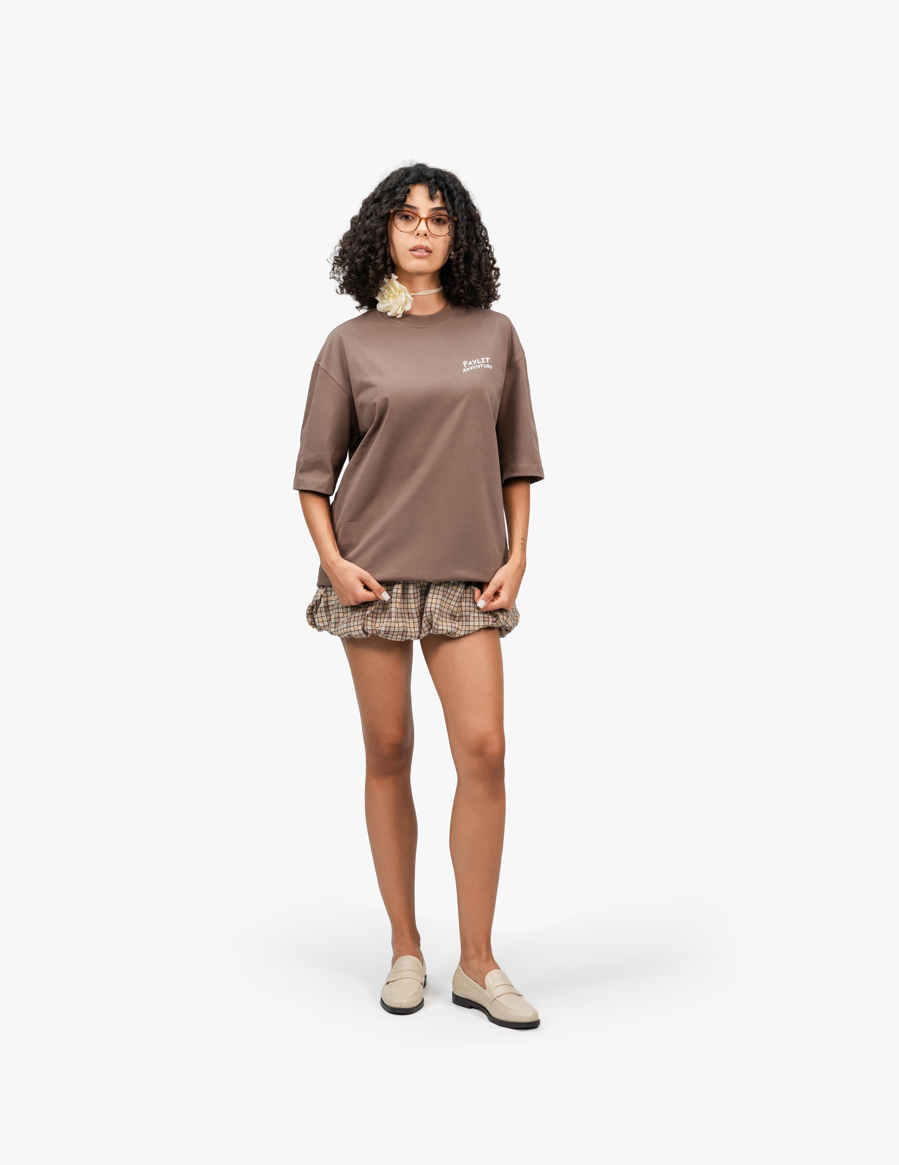 Adventure Kahverengi Oversize Unisex Tişört - Beyaz bir stüdyo fonunun önünde duran, kısa siyah kıvırcık saçlı ve gözlüklü bir kadın. Üzerinde göğüs kısmında beyaz bir logo bulunan kahverengi, geniş kesim bir tişört ve etek ucu fırfırlı, kahverengi ekose desenli bir şort giyiyor. Ayaklarında açık renk loafer ayakkabılar var ve boynunda beyaz bir çiçek detayı görülüyor. - Faylit - 