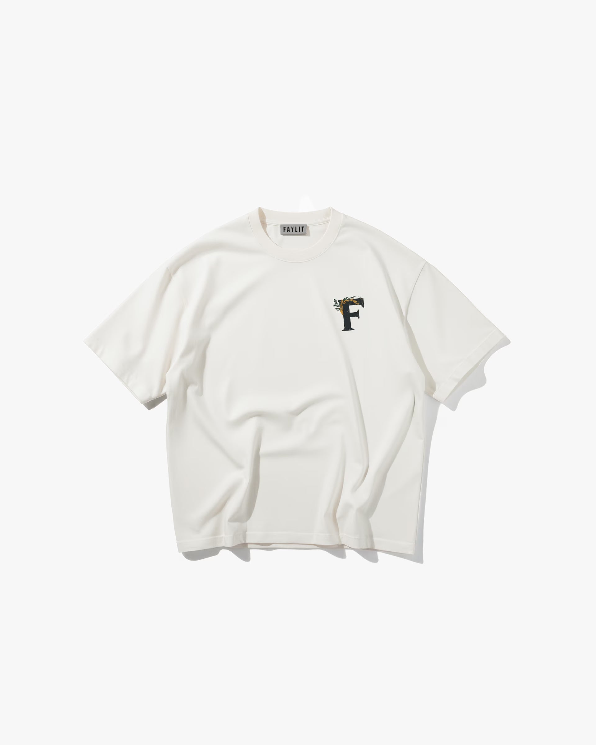 Ancient Ambition Vintage Off White Oversize Tişört Kırık beyaz (off-white) oversize tişörtün ön yüzünün düz çekim ürün fotoğrafı. Tişörtün sol göğsünde, küçük ve detaylı bir logo yer alıyor. Logo, defne yaprakları ile süslenmiş, antik bir görünüme sahip 'F' harfinden oluşuyor. Yeşil ve sarı tonları tasarıma hakimdir.