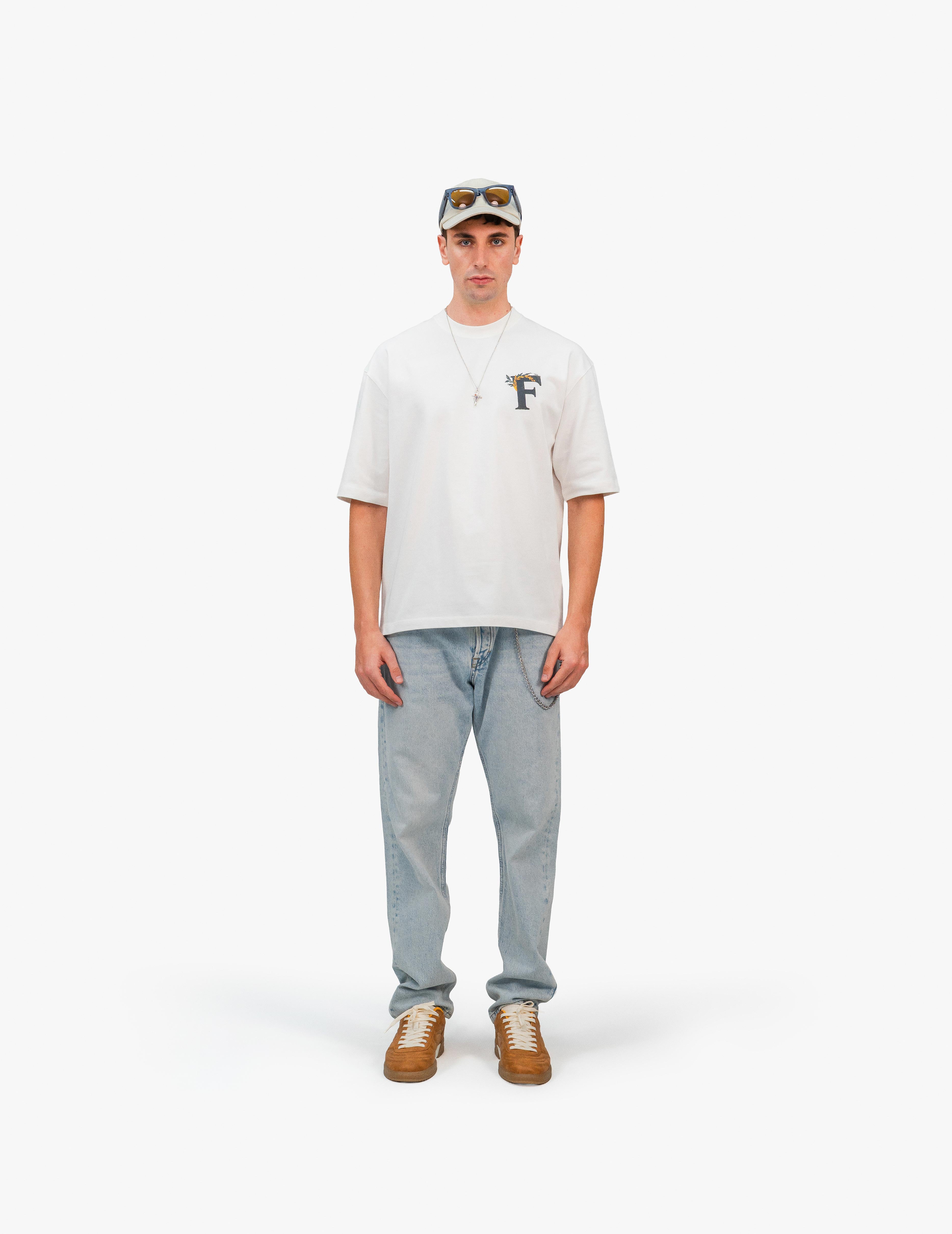 Ancient Ambition Vintage Off White Oversize Tişört, Beyaz bir stüdyo fonunun önünde, doğrudan kameraya bakan, kısa kahverengi saçlı ve şapkalı bir erkek. Başındaki şapka üzerinde güneş gözlüğü var. Üzerinde sol göğüs hizasında "F" harfi içeren renkli bir grafik baskı bulunan beyaz, geniş kesim bir tişört ve açık mavi, bol kesim bir kot pantolon giyiyor. Boynunda gümüş renkli bir kolye var ve ayaklarında kahverengi ayakkabılar bulunuyor. -  Faylit