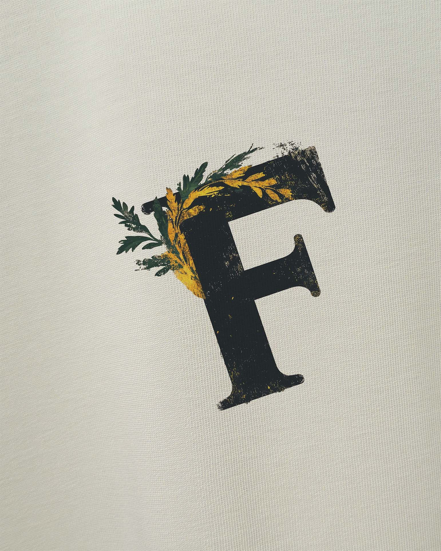 Ancient Ambition Vintage Off White Oversize Tişört Kırık beyaz tişört üzerindeki logo baskısının detaylı yakın çekimi. Eskitme dokulu siyah 'F' harfi, yeşil defne yaprakları ve sarı renkli bitkisel desenlerle süslenmiştir. Tasarım, antik ve botanik öğeleri birleştirerek vintage bir hava yaratıyor. Kumaşın dokusu da görülebilmektedir. -  Faylit