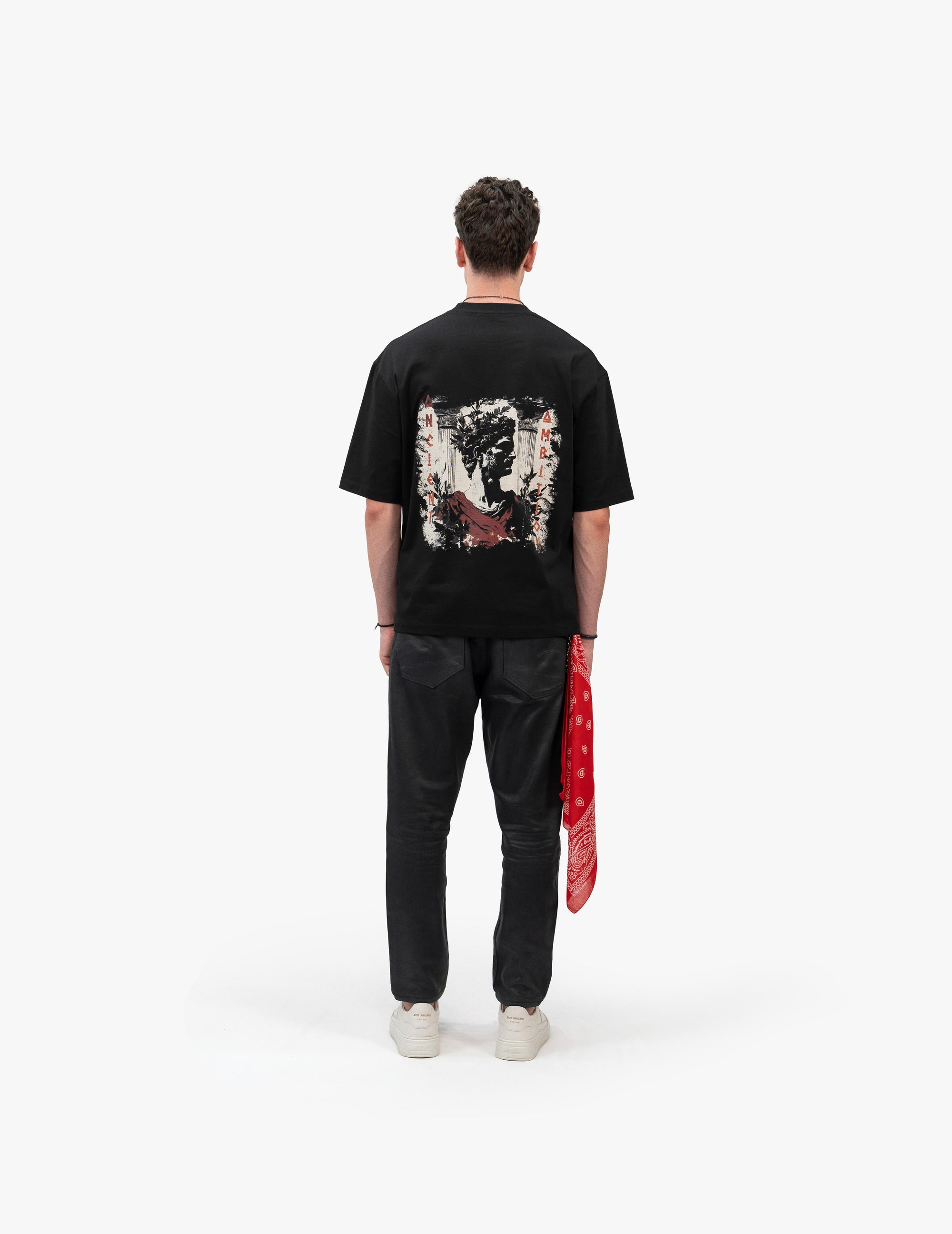 Beyaz fonda, arkası dönük erkek manken, Ancient Ambition Vintage Siyah Oversize Tişört giyiyor. Tişörtün sırtında beyaz, kırmızı ve siyah fırça darbelerinden oluşan soyut bir desen var. Siyah dar pantolon ve beyaz spor ayakkabılarla kombinlenmiş. Sağ arka cebinden kırmızı desenli bir bandana sarkıyor.