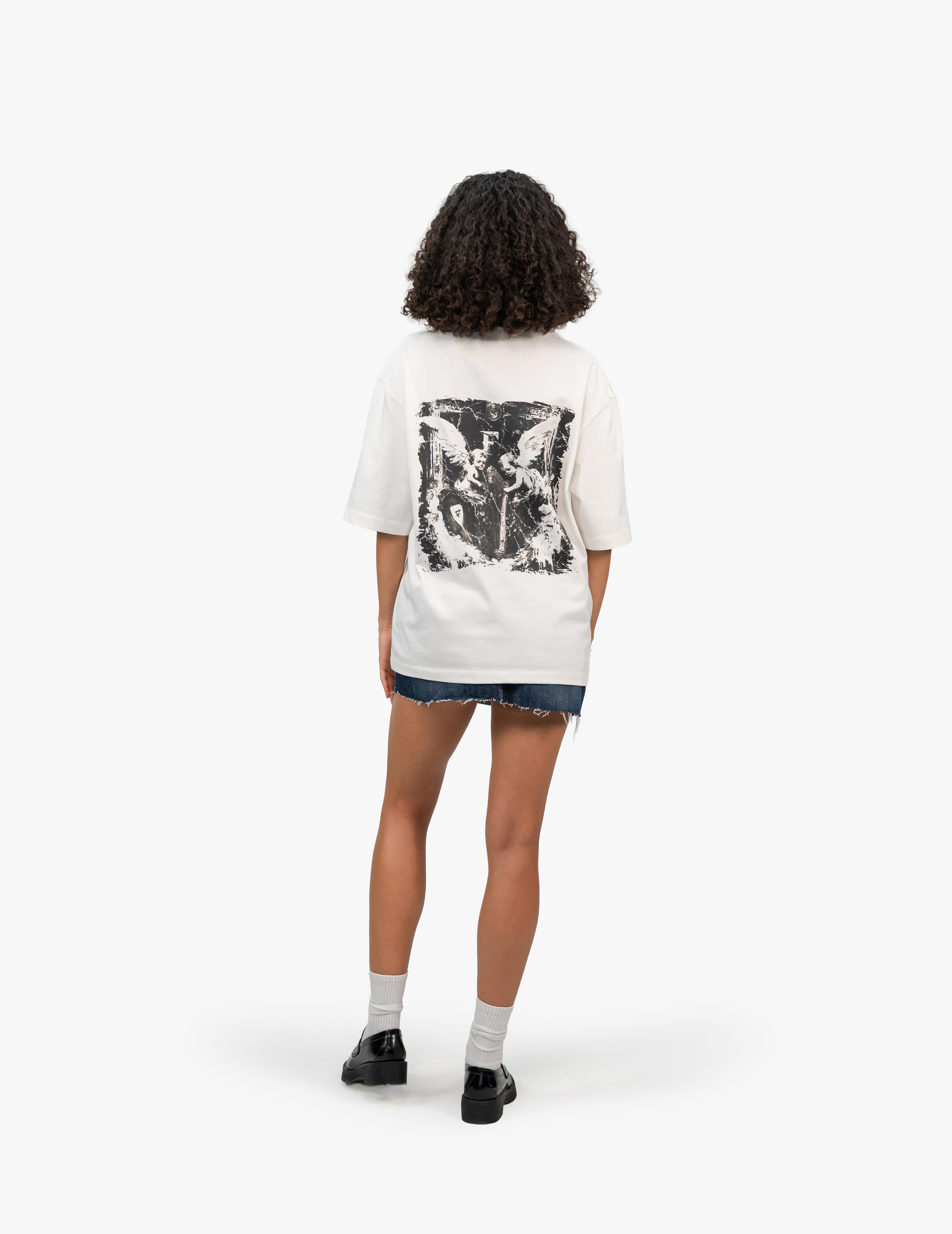 Arp Angels Off White Oversize Tişört, Beyaz bir fonun önünde, arkası dönük bir şekilde duran, kısa siyah kıvırcık saçlı bir kadın. Üzerinde sırt kısmında yıpranmış görünümlü, melek figürlü siyah beyaz bir grafik baskı bulunan beyaz, geniş kesim bir tişört ve lacivert, yırtık etekli bir mini kot etek giyiyor. Ayaklarında beyaz çoraplar ve siyah, rugan loafer ayakkabılar bulunuyor. -  Faylit