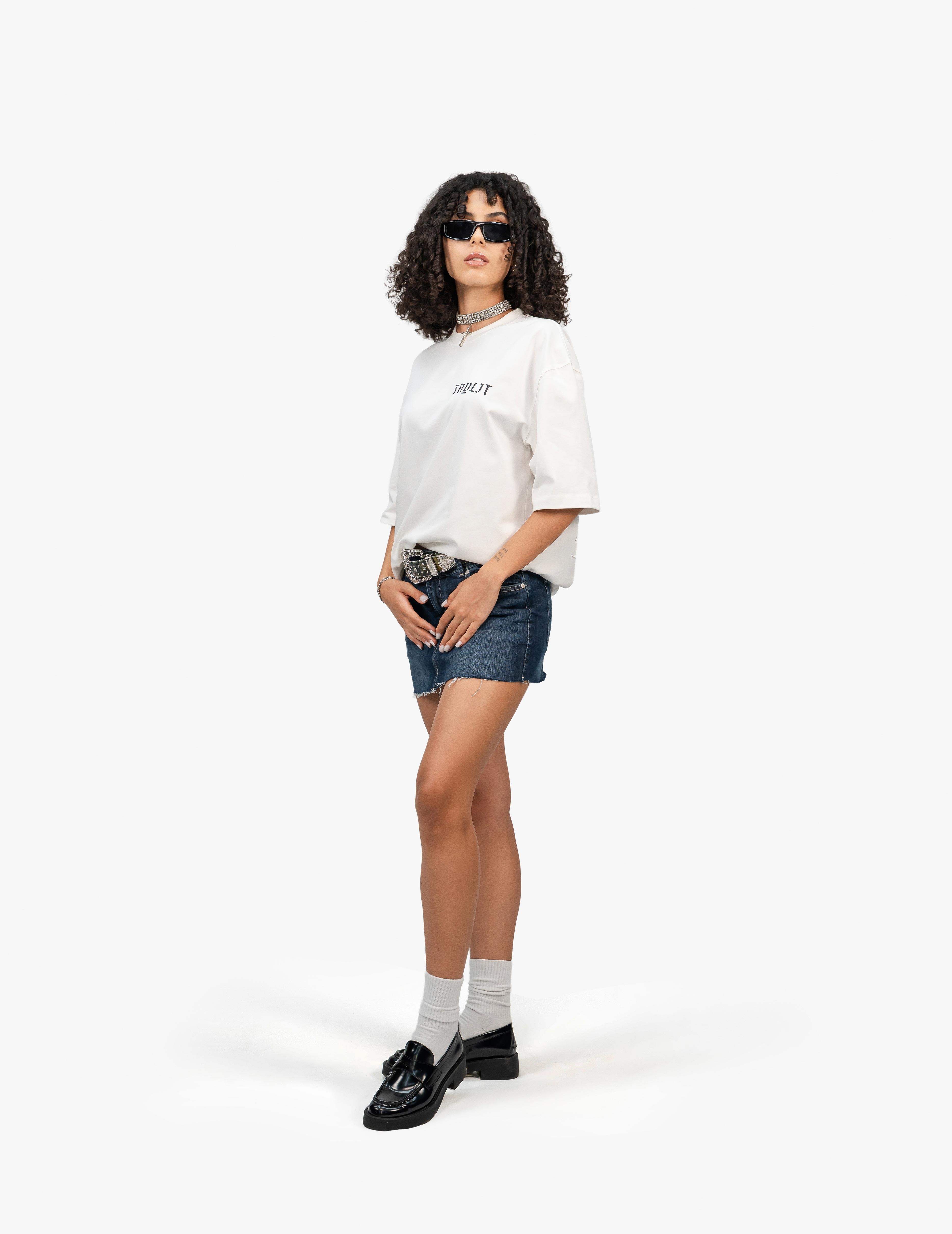 Arp Angels Off White Oversize Tişört, Beyaz bir fonun önünde, hafifçe sola dönük duran, kısa siyah kıvırcık saçlı bir kadın. Üzerinde göğüs kısmında "Faylit" yazısı bulunan beyaz, geniş kesim bir tişört ve lacivert, yırtık etekli bir mini kot etek giyiyor. Eteğin üzerinde metal tokalı bir kemer var. Elleri cebinde. Ayaklarında beyaz çoraplar ve siyah, rugan loafer ayakkabılar bulunuyor. Boynunda gümüş renkli bir kolye ve gözünde siyah çerçeveli güneş gözlüğü takıyor. -  Faylit