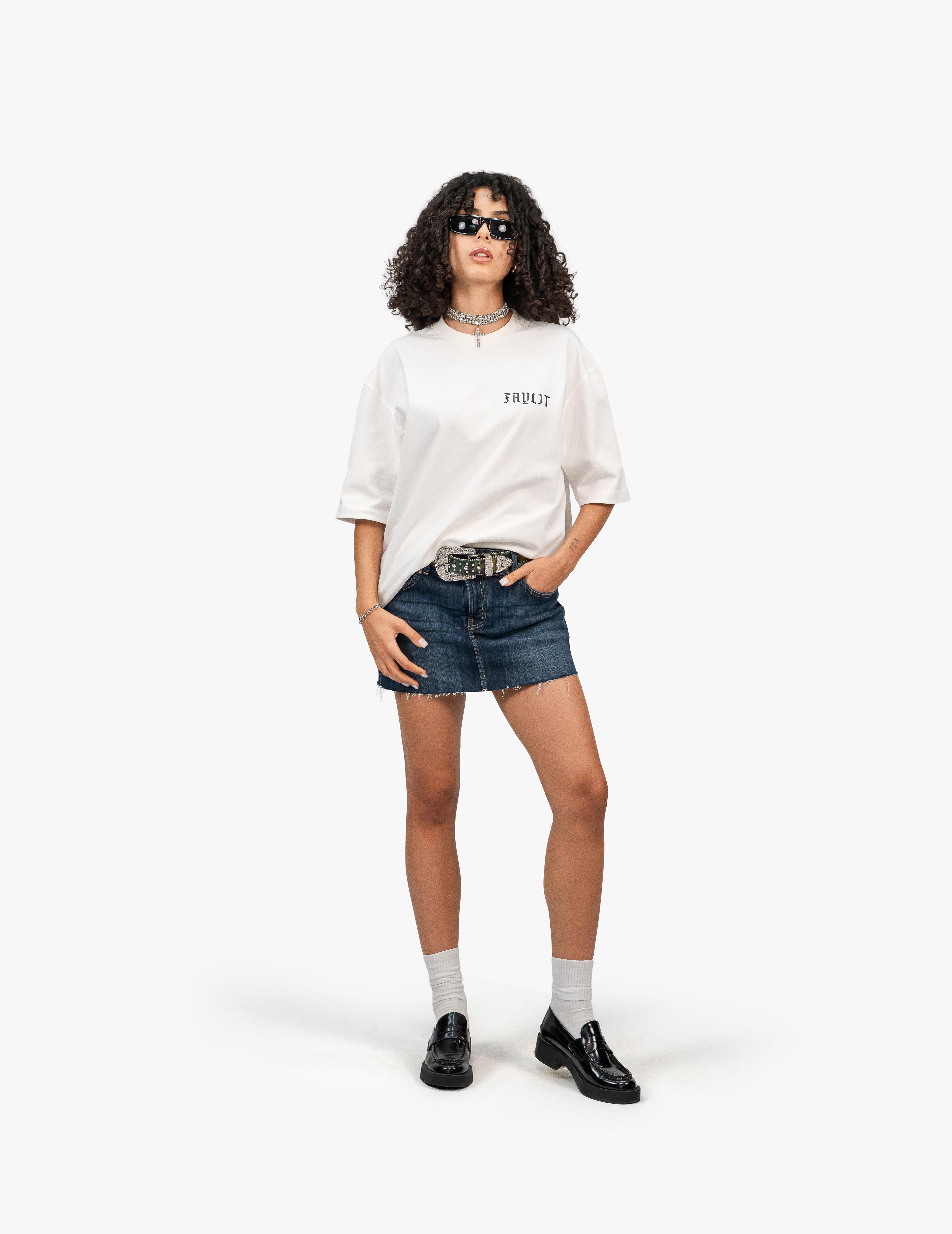 Arp Angels Off White Oversize Tişört, Beyaz bir fonun önünde, doğrudan kameraya bakan, kısa siyah kıvırcık saçlı bir kadın. Üzerinde göğüs kısmında "Faylit" yazısı bulunan beyaz, geniş kesim bir tişört ve lacivert, yırtık etekli bir mini kot etek giyiyor. Eteğin üzerinde metal tokalı bir kemer var. Elleri cebinde. Ayaklarında beyaz çoraplar ve siyah, rugan loafer ayakkabılar bulunuyor. Boynunda gümüş renkli bir kolye ve gözünde siyah çerçeveli güneş gözlüğü takıyor. -  Faylit