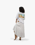 Beach Vibes Optik Beyaz Oversize Unisex Tişört, Beyaz bir fonun önünde, arkası dönük ve hafifçe sola dönük duran, kısa siyah kıvırcık saçlı bir kadın. Üzerinde sırt kısmında palmiyeler, bir papağan ve bir golf arabası içeren plaj temalı renkli bir grafik baskı bulunan beyaz, geniş kesim bir tişört ve uzun, beyaz, kat kat bir etek giyiyor. Ayaklarında sivri burunlu, açık kahverengi, topuklu botlar bulunuyor. -  Faylit