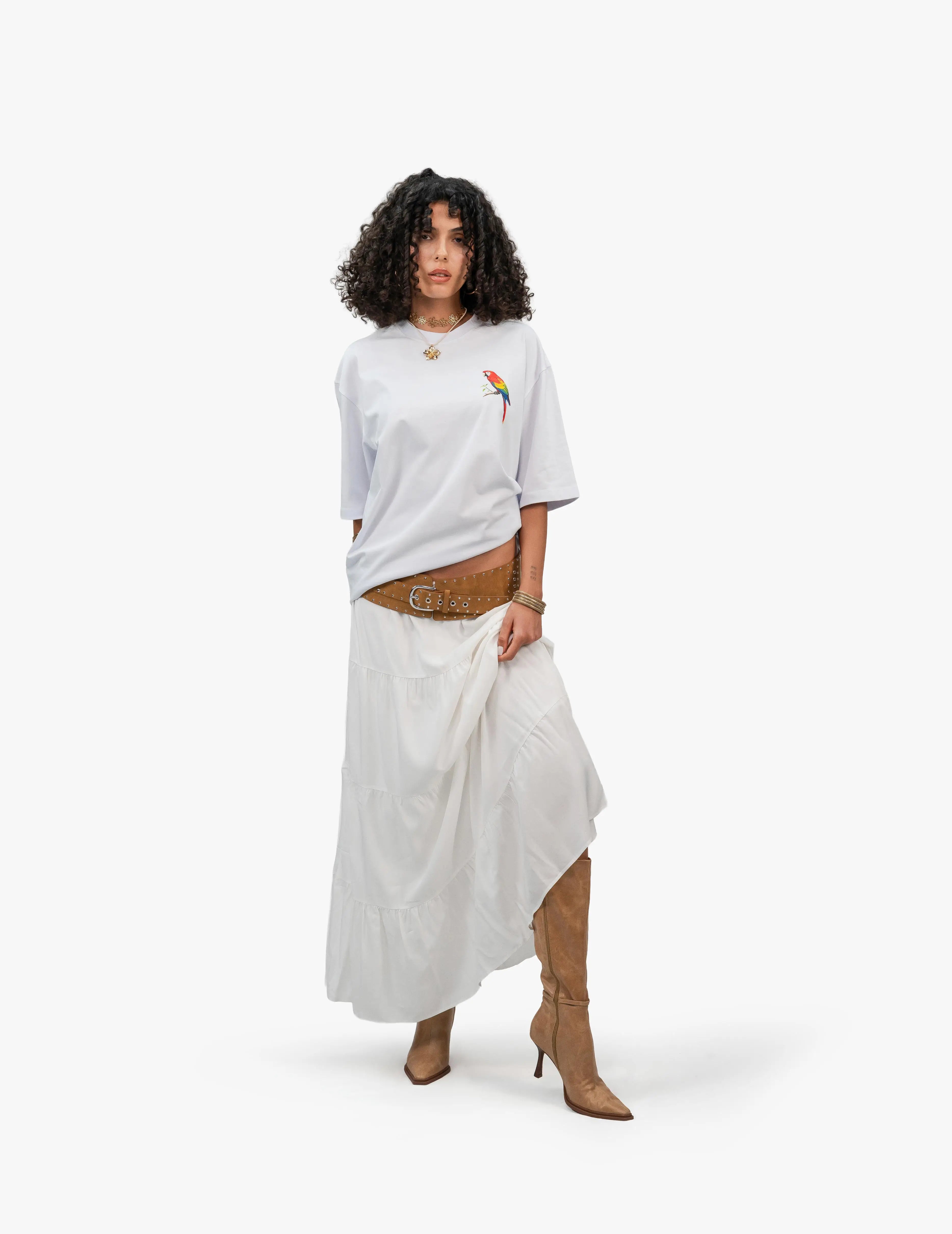 Beach Vibes Optik Beyaz Oversize Unisex Tişört, Beyaz bir fonun önünde, doğrudan kameraya bakan, kısa siyah kıvırcık saçlı bir kadın. Üzerinde sağ göğüs hizasında renkli bir papağan figürü bulunan beyaz, geniş kesim bir tişört ve uzun, beyaz, kat kat bir etek giyiyor. Eteğin üzerinde kalın, kahverengi, metal detaylı bir kemer var. Ayaklarında sivri burunlu, açık kahverengi, topuklu botlar bulunuyor. Boynunda altın renkli, çiçek desenli bir gerdanlık takıyor. -  Faylit