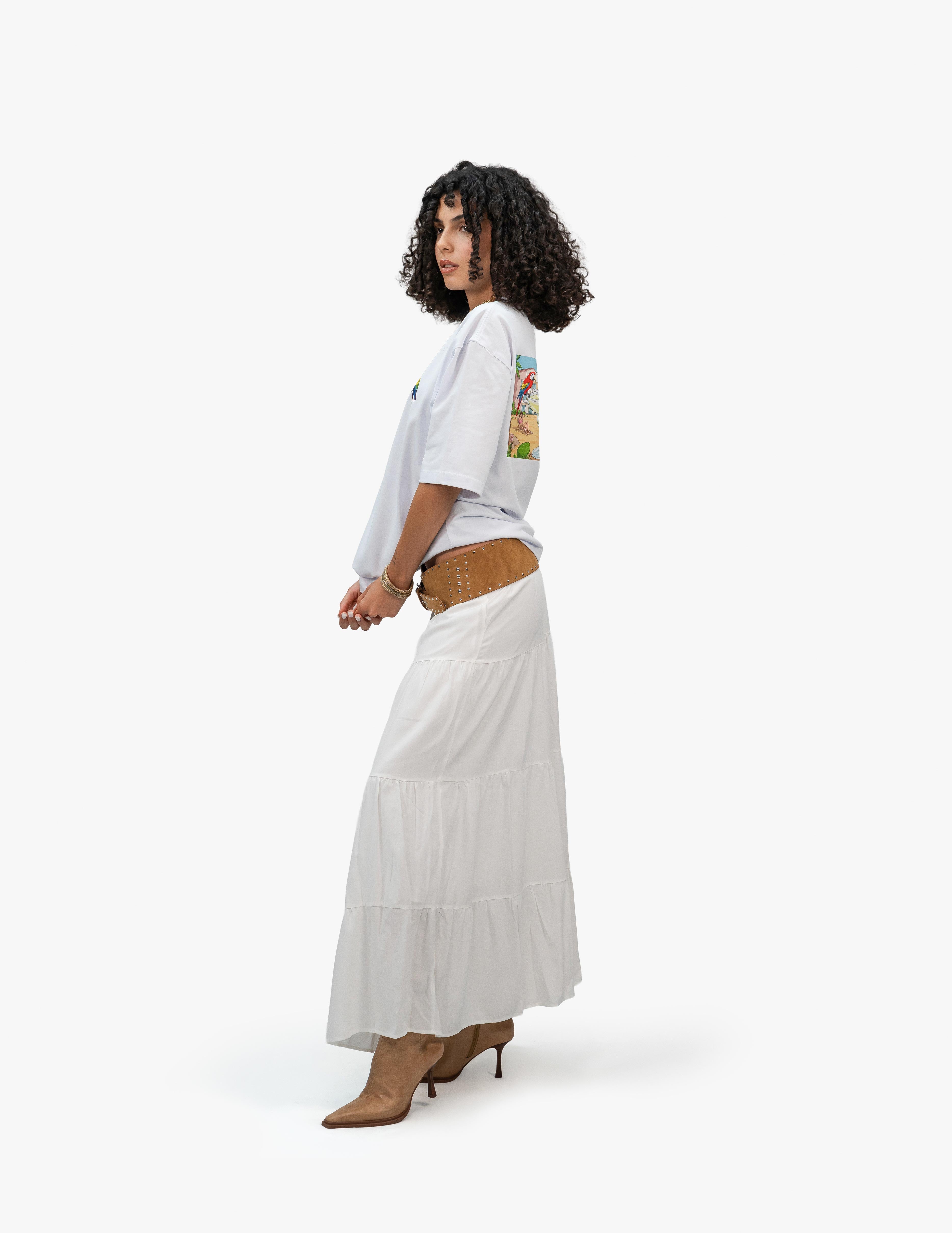 Beach Vibes Optik Beyaz Oversize Unisex Tişört, Beyaz bir fonun önünde, vücudu ve başı sağa dönük duran, kısa siyah kıvırcık saçlı bir kadın. Üzerinde sırt kısmında plaj temalı renkli bir grafik baskı bulunan beyaz, geniş kesim bir tişört ve uzun, beyaz, kat kat bir etek giyiyor. Eteğin üzerinde kalın, kahverengi, metal detaylı bir kemer var. Elleri tişörtünün etek kısmının önünde birleşmiş. Ayaklarında sivri burunlu, açık kahverengi, topuklu botlar bulunuyor. -  Faylit