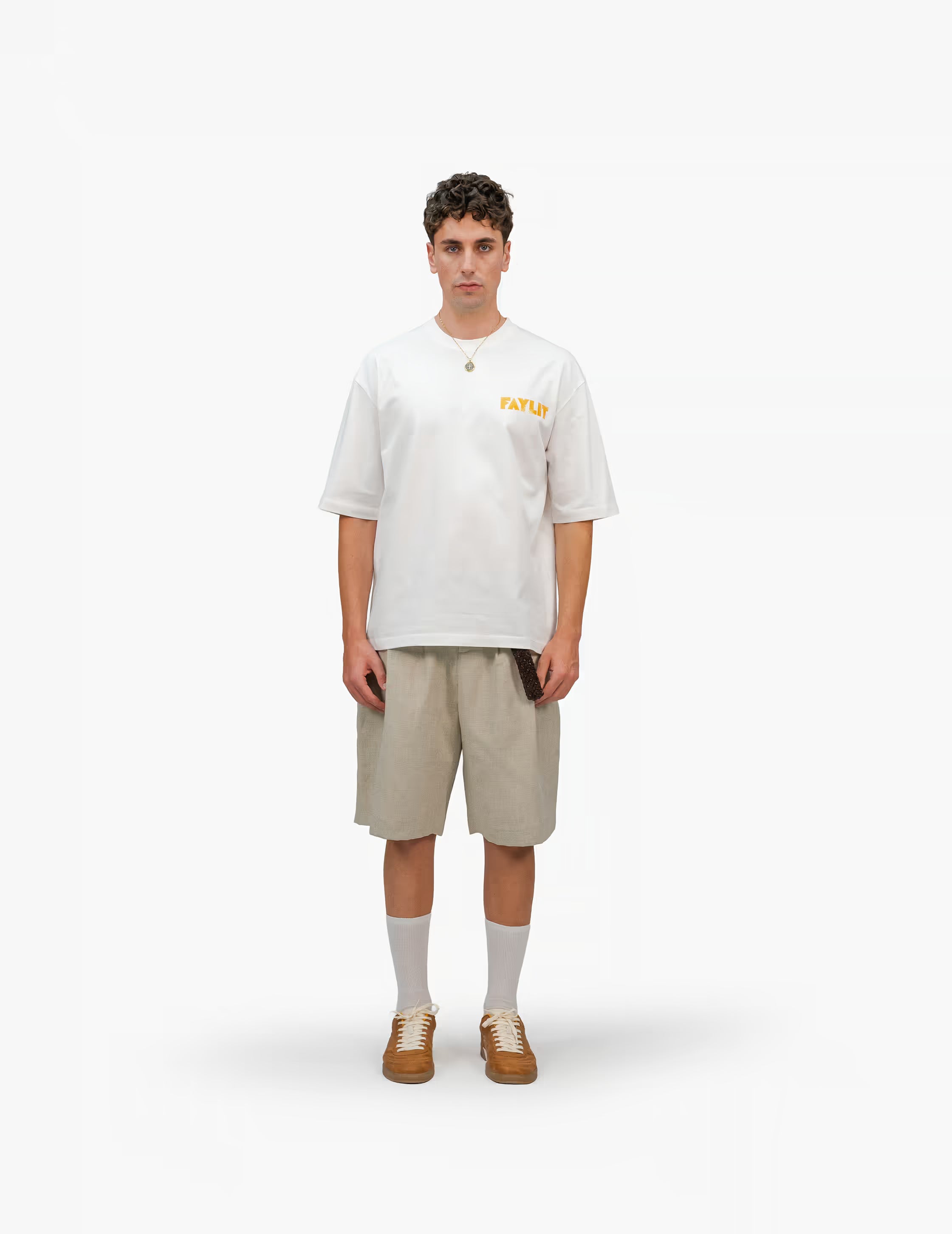 Erkek modelin üzerindeki FAYLIT oversize beyaz t-shirt, göğüste turuncu FAYLIT logosu, şort ve spor ayakkabı ile kombinlenmiş, rahat ve şık günlük giyim.