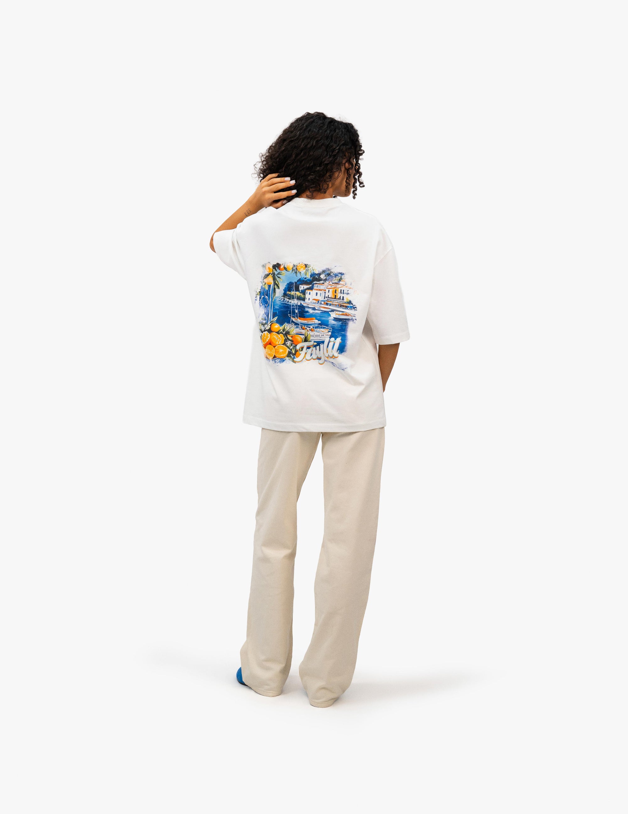Coast Beach Vintage Off White Oversize Tişört- Beyaz stüdyo fonunda, arkası dönük kıvırcık saçlı kadın manken, sırtında renkli bir İtalyan Riviera manzara baskısı olan beyaz oversize bir tişört ve bej rengi bol pantolon giyiyor. Manzara baskısında evler, deniz, meyveler ve "Faylit" yazısı görülüyor. -  Faylit