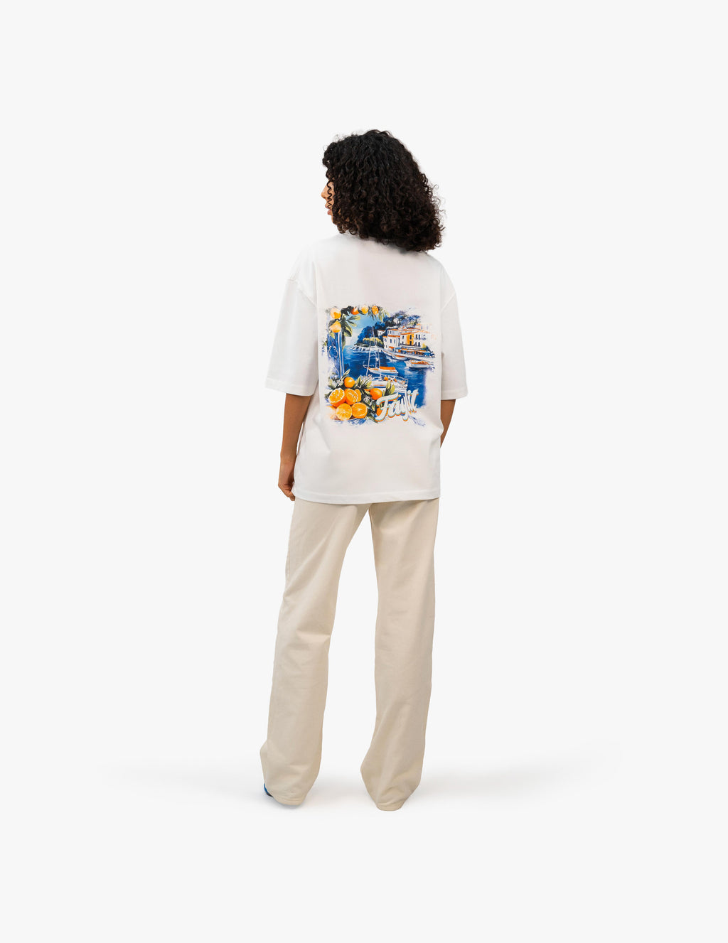 Coast Beach Vintage Off White Oversize Tişört Beyaz stüdyo fonunda, arkası dönük kıvırcık saçlı kadın manken, sırtında renkli bir İtalyan Riviera manzara baskısı olan beyaz oversize bir tişört ve bej rengi bol pantolon giyiyor. Manzara baskısında canlı renkler ve detaylı bir sahil kasabası tasviri bulunuyor. -  Faylit
