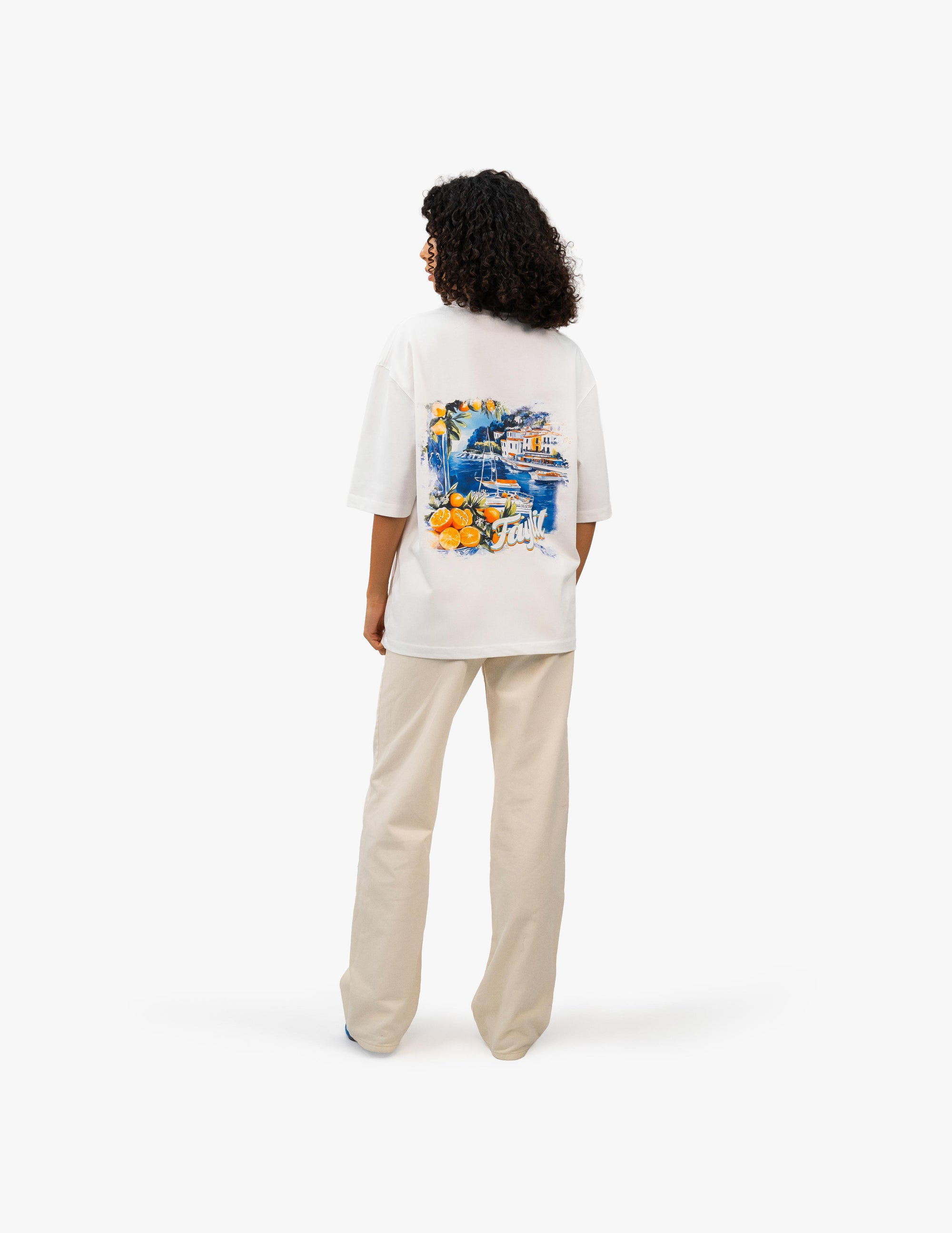 Coast Beach Vintage Off White Oversize Tişört Beyaz stüdyo fonunda, arkası dönük kıvırcık saçlı kadın manken, sırtında renkli bir İtalyan Riviera manzara baskısı olan beyaz oversize bir tişört ve bej rengi bol pantolon giyiyor. Manzara baskısında canlı renkler ve detaylı bir sahil kasabası tasviri bulunuyor. -  Faylit