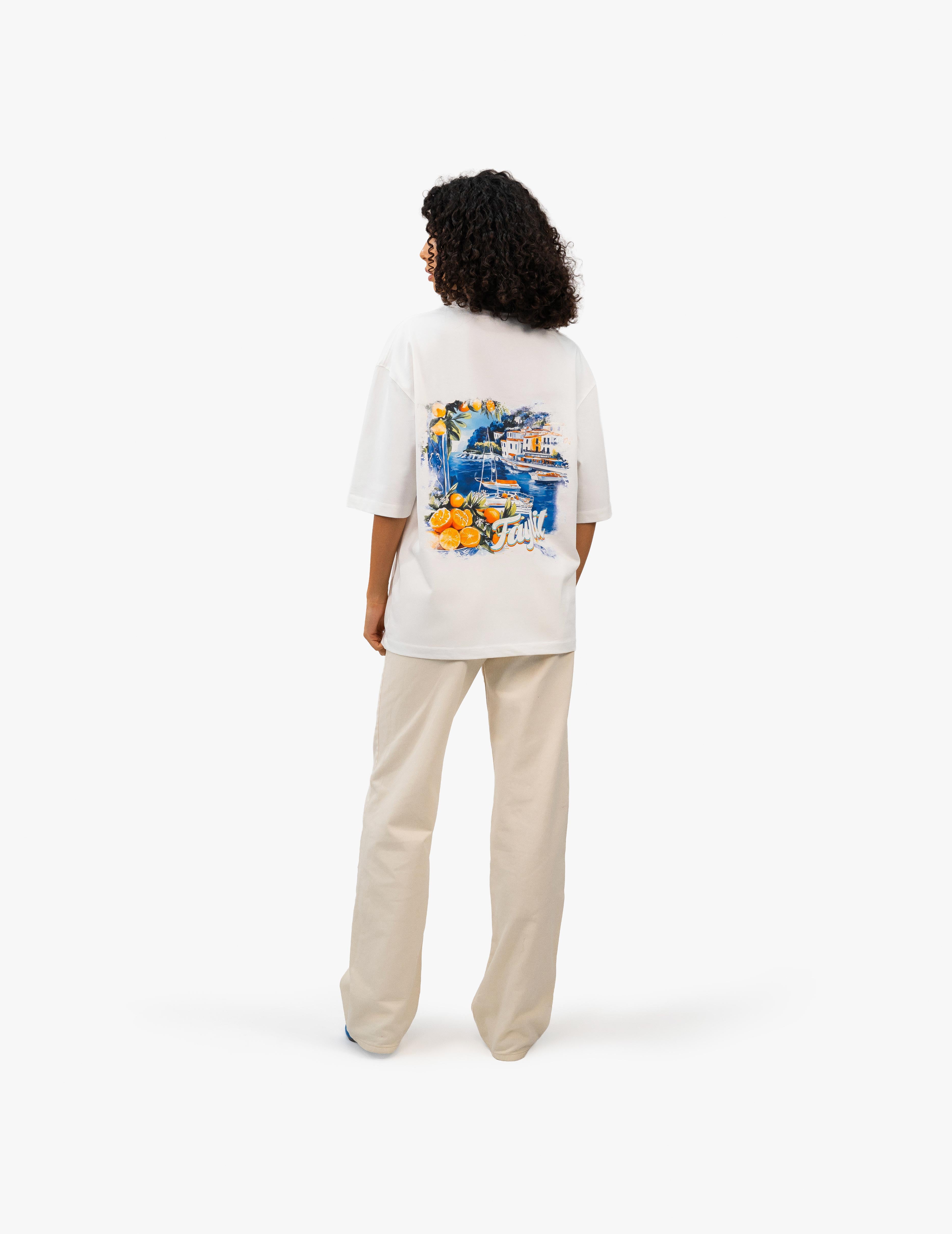 Coast Beach Vintage Off White Oversize Tişört Beyaz stüdyo fonunda, arkası dönük kıvırcık saçlı kadın manken, sırtında renkli bir İtalyan Riviera manzara baskısı olan beyaz oversize bir tişört ve bej rengi bol pantolon giyiyor. Manzara baskısında canlı renkler ve detaylı bir sahil kasabası tasviri bulunuyor. -  Faylit