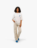 Coast Beach Vintage Off White Oversize Tişört - Beyaz stüdyo fonunda, kıvırcık saçlı bir kadın manken, sol göğsünde mavi, stilize bir "F" harfi bulunan beyaz oversize bir tişört ve bej rengi bol pantolon giyiyor. Rahat ve modern bir görünüm sergiliyor.-  Faylit