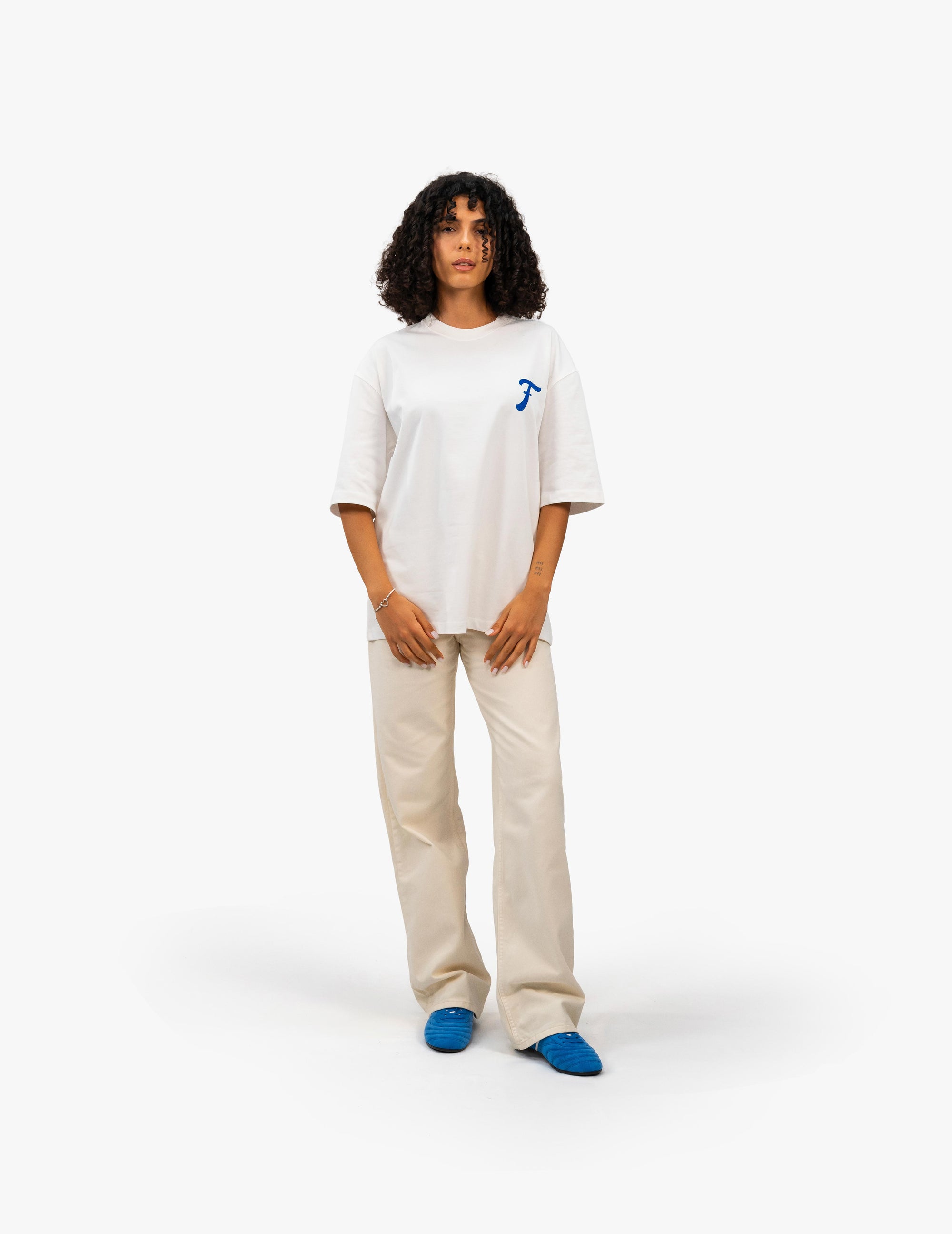 Coast Beach Vintage Off White Oversize Tişört -Beyaz stüdyo fonunda, kıvırcık saçlı bir kadın manken, sol göğsünde mavi, stilize bir "F" harfi bulunan beyaz oversize bir tişört ve bej rengi bol pantolon giyiyor. Kendine güvenli bir duruş sergiliyor. -  Faylit