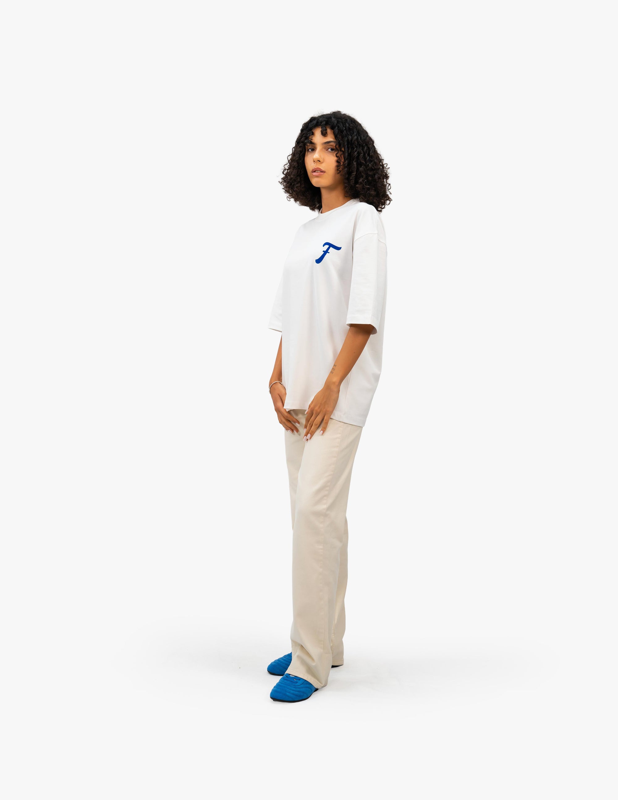 Coast Beach Vintage Off White Oversize Tişört Beyaz stüdyo fonunda, yana dönük kıvırcık saçlı bir kadın manken, sol göğsünde mavi, stilize bir "F" harfi bulunan beyaz oversize bir tişört ve bej rengi bol pantolon giyiyor. Gündelik ve şık bir tarzı yansıtıyor. -  Faylit