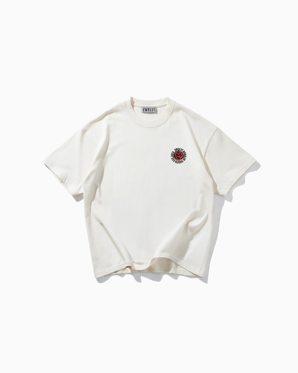 Beyaz bir arka plan üzerinde düz serilmiş, oversize ve rahat kesim krem rengi bir tişörtün ön yüzü. Tişörtün göğsünün sol üst kısmında, kırmızı renkte dairesel bir logo bulunuyor. Logonun içinde 'FAYLIT' markasının adının stilize edilmiş bir versiyonu ve 'BROKEN HEARTS CLUB' gibi küçük yazılar yer alıyor. Tişörtün yakasında da 'FAYLIT' etiketi görünüyor. Sade ve minimalist ön tasarımı, arkasındaki detaylı baskıya kontrast oluşturuyor.