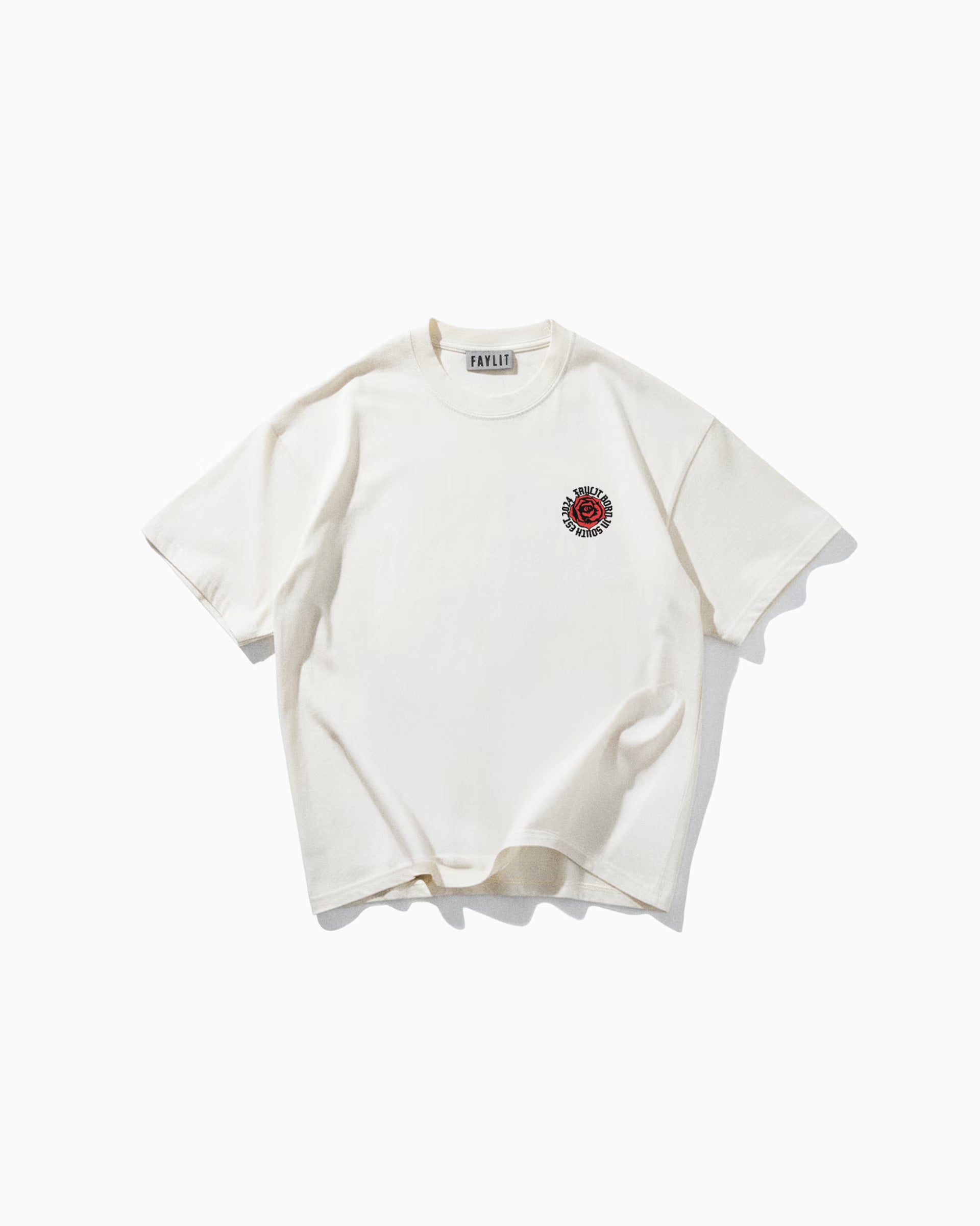 Beyaz bir arka plan üzerinde düz serilmiş, oversize ve rahat kesim krem rengi bir tişörtün ön yüzü. Tişörtün göğsünün sol üst kısmında, kırmızı renkte dairesel bir logo bulunuyor. Logonun içinde 'FAYLIT' markasının adının stilize edilmiş bir versiyonu ve 'BROKEN HEARTS CLUB' gibi küçük yazılar yer alıyor. Tişörtün yakasında da 'FAYLIT' etiketi görünüyor. Sade ve minimalist ön tasarımı, arkasındaki detaylı baskıya kontrast oluşturuyor.