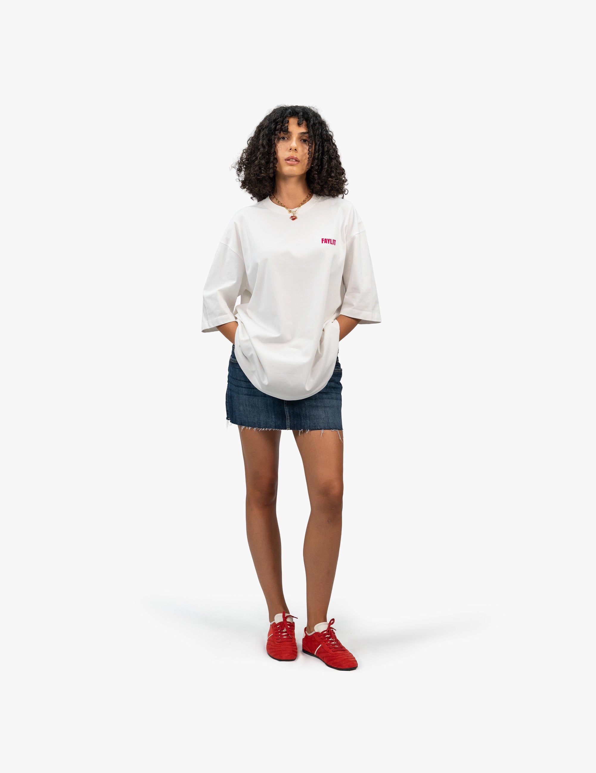 FDNA_07 Oversize Tişört - Beyaz stüdyo fonunda, elleri cebinde poz veren kıvırcık saçlı kadın manken. Üzerinde, sol göğsünde pembe "FAYLIT" logosu bulunan beyaz oversize bir tişört, yıpranmış paçalı kot mini etek ve canlı kırmızı renkte spor ayakkabılar giyiyor. -  Faylit