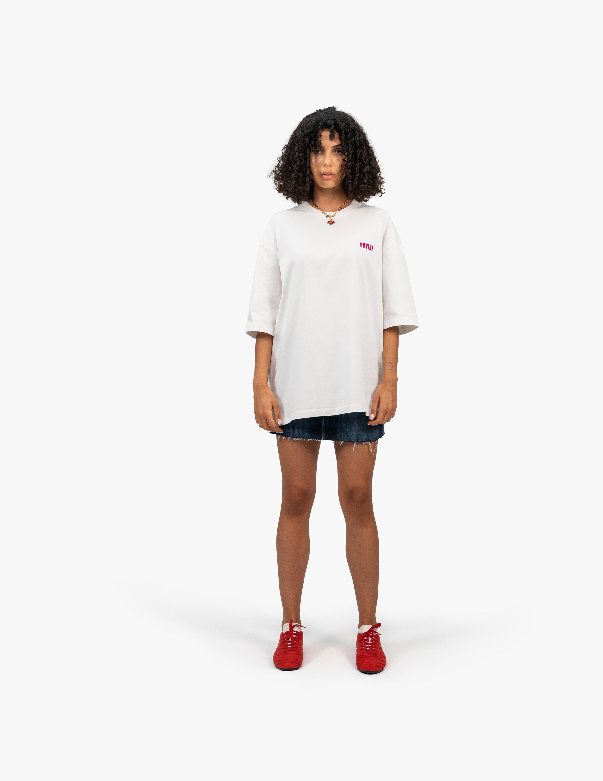 FDNA_07 Oversize Tişört - Beyaz stüdyo fonunda, tam karşıdan poz veren kıvırcık saçlı kadın manken. Üzerinde, sol göğsünde pembe "FAYLIT" logosu bulunan beyaz oversize bir tişört, yıpranmış görünümlü kot şort ve canlı kırmızı renkte spor ayakkabılar var. Modern ve gündelik bir yaz stili sergiliyor. -  Faylit