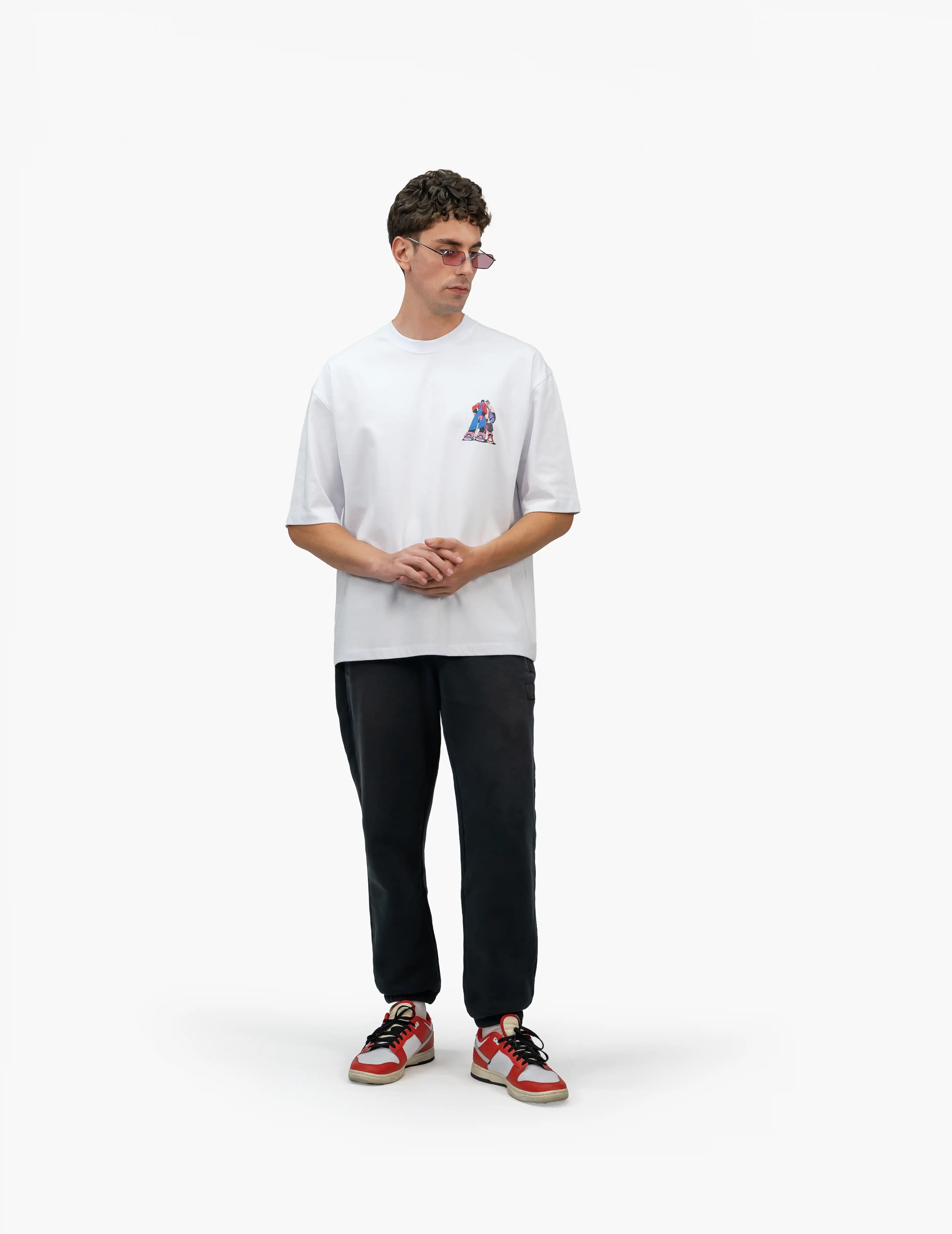 Faylit Style Beyaz Oversize Tişört, Beyaz bir stüdyo fonunun önünde, hafifçe sağa dönük duran, kısa kahverengi saçlı ve güneş gözlüklü bir erkek. Üzerinde sağ göğüs hizasında renkli bir grafik baskı bulunan beyaz, geniş kesim bir tişört ve siyah, bol kesim bir eşofman altı giyiyor. Ellerini göğsünün önünde birleştirmiş. Ayaklarında kırmızı, beyaz ve siyah renkli spor ayakkabılar bulunuyor. -  Faylit