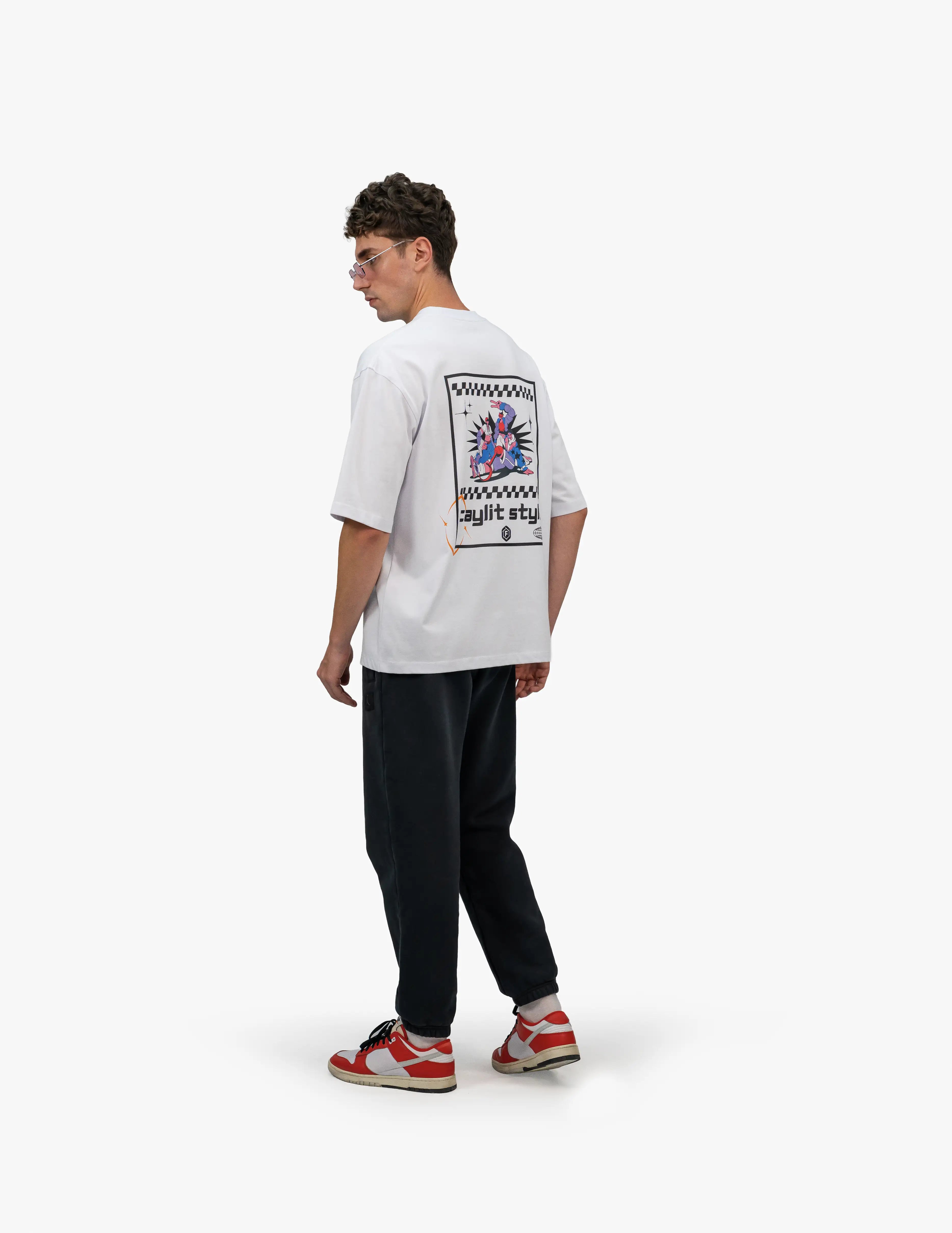 Faylit Style Beyaz Oversize Tişört, Beyaz bir stüdyo fonunun önünde, arkası dönük ve hafifçe sola dönük bir şekilde duran, kısa kahverengi saçlı ve güneş gözlüklü bir erkek. Başını sağa çevirmiş şekilde bakıyor. Üzerinde sırtında renkli ve "Faylit Style" yazısı içeren, dama tahtası desenli ve çizgili bir grafik baskı bulunan beyaz, geniş kesim bir tişört ve siyah, bol kesim bir eşofman altı giyiyor. Ayaklarında kırmızı, beyaz ve siyah renkli spor ayakkabılar bulunuyor. -  Faylit