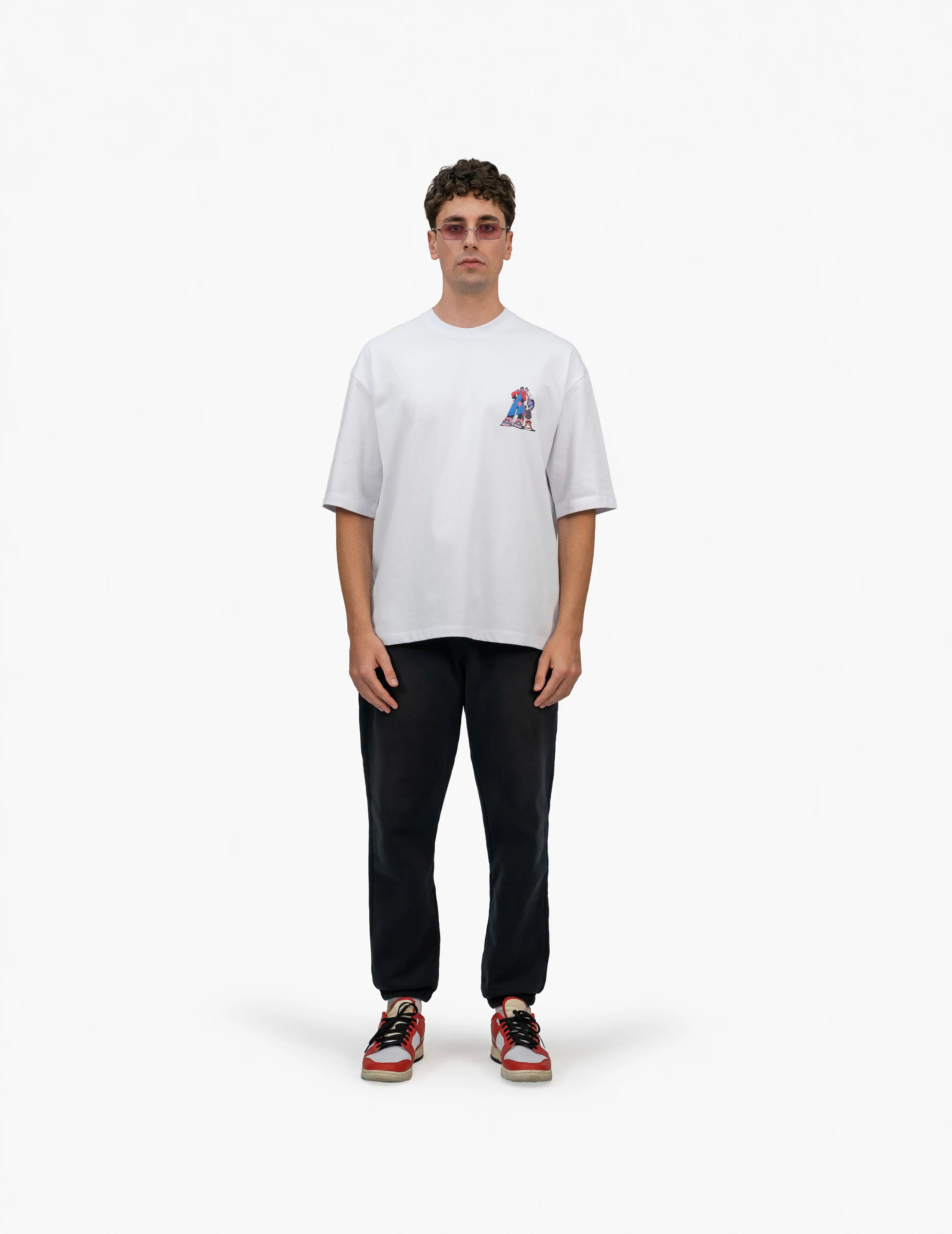 Faylit Style Beyaz Oversize Tişört, Beyaz bir stüdyo fonunun önünde, doğrudan kameraya bakan, kısa kahverengi saçlı ve güneş gözlüklü bir erkek. Üzerinde sağ göğüs hizasında renkli bir grafik baskı bulunan beyaz, geniş kesim bir tişört ve siyah, bol kesim bir eşofman altı giyiyor. Ayaklarında kırmızı, beyaz ve siyah renkli spor ayakkabılar bulunuyor. -  Faylit