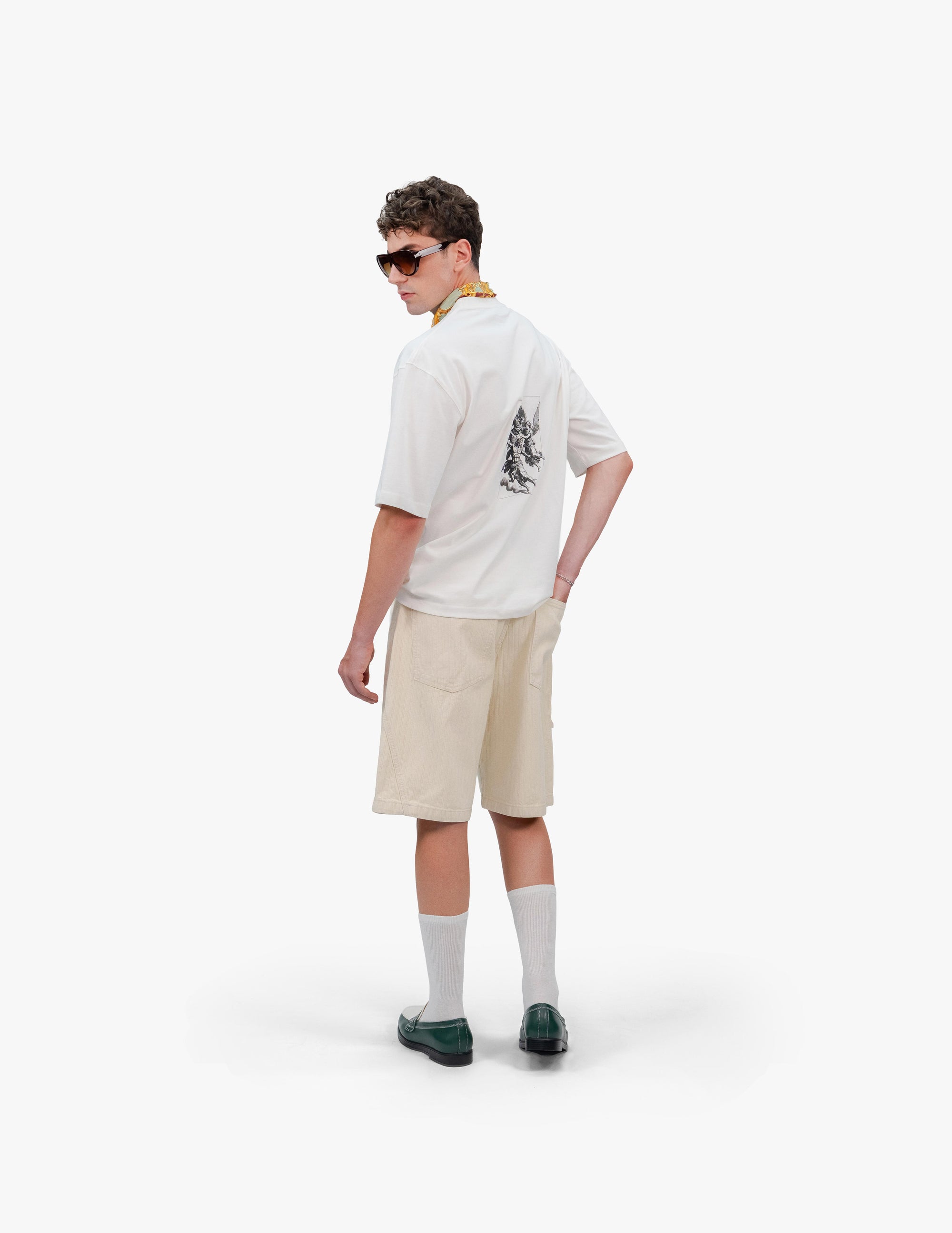 Gods Off White Oversize Tişört Beyaz stüdyo fonunda, arkası dönük duran ve omzunun üzerinden geriye bakan erkek manken. Sırtında, dikey ve gravür stilinde meleklerin mücadelesini tasvir eden bir baskı bulunan beyaz oversize tişört giyiyor. Kombini, bej rengi şort, beyaz çorap ve siyah makosen ayakkabılarla tamamlanmış. Boynundaki yeşil fular ve taktığı güneş gözlüğü de görülüyor. -  Faylit
