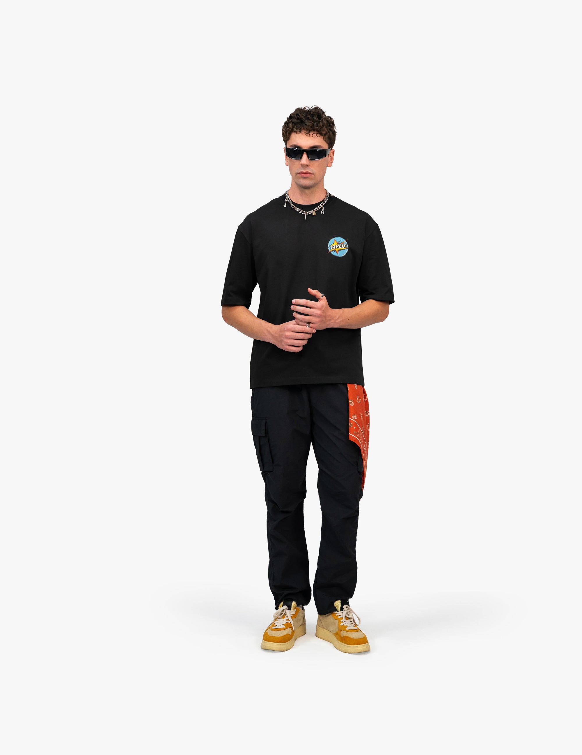 Lambo Premium Vintage Siyah Tişört - Beyaz stüdyo fonunda poz veren erkek manken. Üzerinde, sol göğsünde daire şeklinde renkli bir manzara logosu bulunan siyah oversize tişört, siyah kargo pantolon ve hardal sarısı spor ayakkabılar var. Kombin, modern güneş gözlükleri, katmanlı kolyeler ve cebindeki kırmızı bandana ile tamamlanıyor. -  Faylit