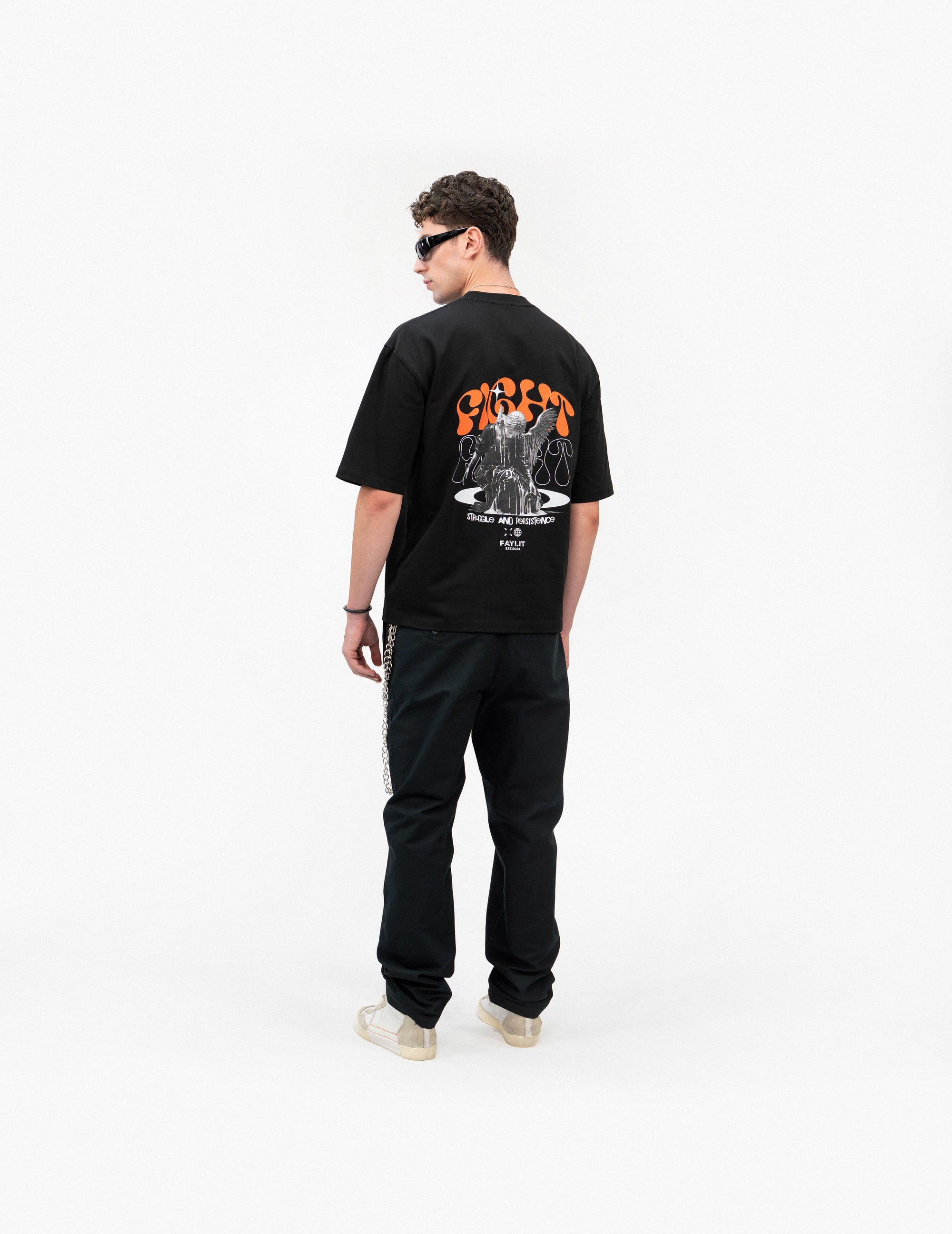 Persistence Retro Grafik Siyah Oversize Tişört - Beyaz stüdyo fonunda, arkası dönük duran ve omzunun üzerinden yana bakan erkek manken. Sırtında, turuncu "FIGHT" yazılı ve mücadele eden melek figürlü büyük bir baskı bulunan siyah oversize bir tişört giyiyor. Kombini, siyah pantolon ve açık renkli, bilekli spor ayakkabılarla tamamlanmış. Pantolonundan sarkan gümüş rengi zincir detayı dikkat çekiyor. -  Faylit