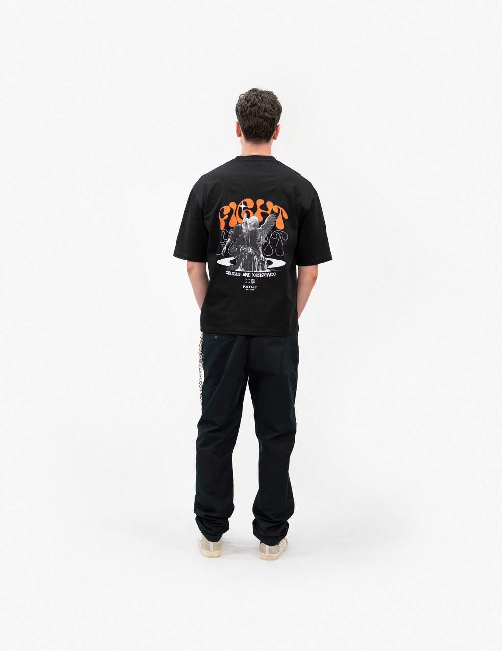 Persistence Retro Grafik Siyah Oversize Tişört - Beyaz stüdyo fonunda, arkası dönük olarak poz veren erkek manken. Sırtında, turuncu "FIGHT" yazılı ve mücadele eden melek figürlü büyük bir baskı bulunan siyah oversize bir tişört giyiyor. Kombini, siyah pantolon ve açık renkli, bilekli spor ayakkabılarla tamamlanmış. Sol cebinden gümüş rengi bir pantolon zinciri sarkmaktadır. -  Faylit