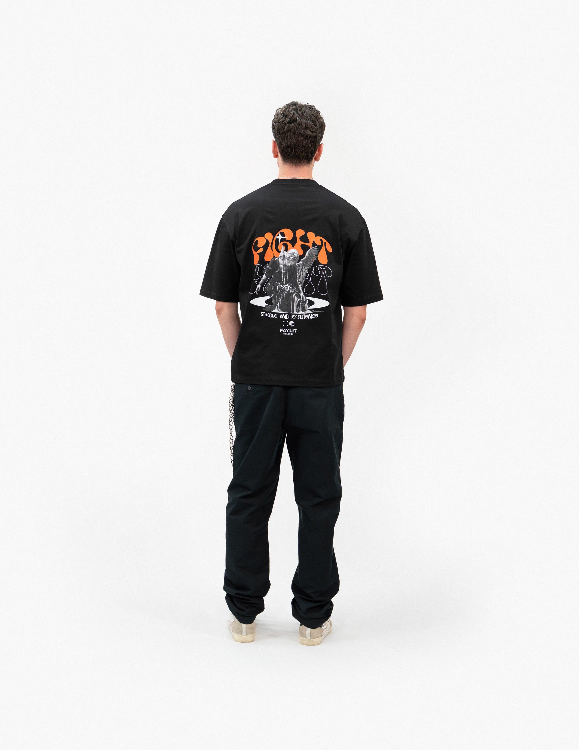 Persistence Retro Grafik Siyah Oversize Tişört - Beyaz stüdyo fonunda, arkası dönük olarak poz veren erkek manken. Sırtında, turuncu "FIGHT" yazılı ve mücadele eden melek figürlü büyük bir baskı bulunan siyah oversize bir tişört giyiyor. Kombini, siyah pantolon ve açık renkli, bilekli spor ayakkabılarla tamamlanmış. Sol cebinden gümüş rengi bir pantolon zinciri sarkmaktadır. -  Faylit