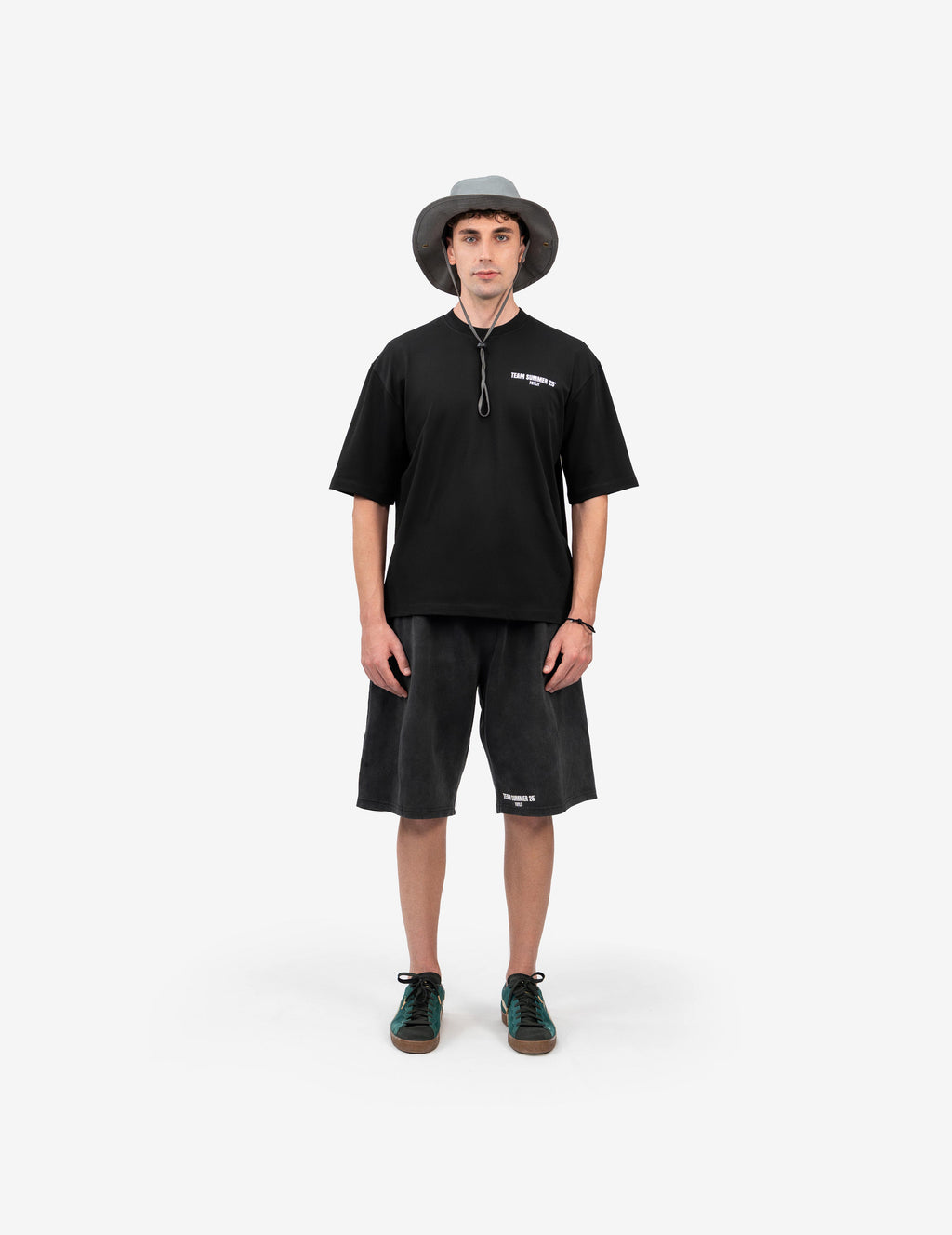 Team Summer 25 Siyah Oversize Tişört- Beyaz stüdyo fonunda, tam karşıdan poz veren erkek manken, outdoor ve sokak stilini birleştiren bir kombin sergiliyor. Başında ipli, gri bir balıkçı şapkası (bucket hat) takıyor. Üzerinde, göğsünde beyaz yazı baskısı olan siyah oversize bir tişört ve eskitme görünümlü, yine beyaz baskılı antrasit rengi bol bir şort var. Kombinini, yeşil detaylı ve kahverengi tabanlı spor ayakkabılarla tamamlamış. -  Faylit