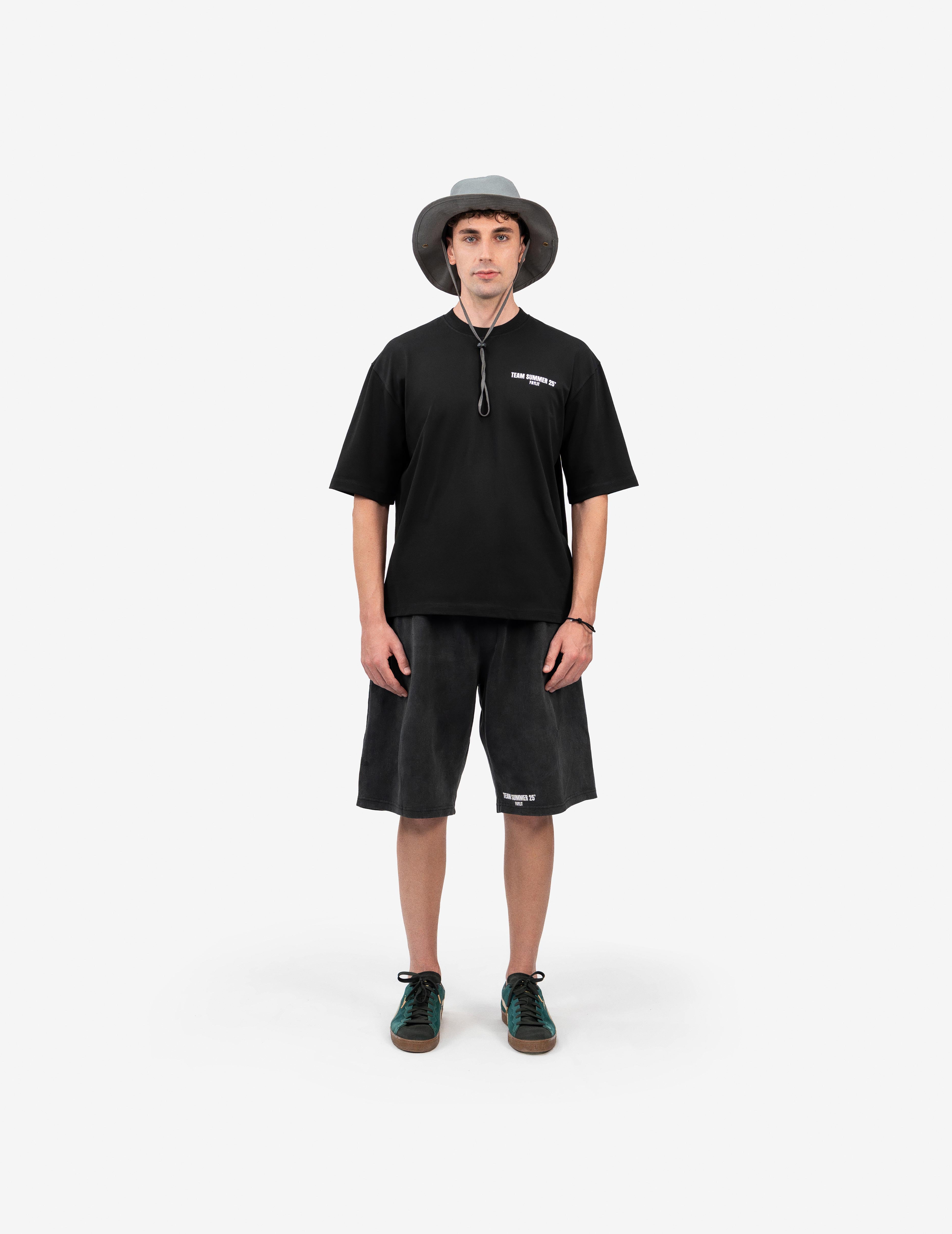 Team Summer 25 Siyah Oversize Tişört- Beyaz stüdyo fonunda, tam karşıdan poz veren erkek manken, outdoor ve sokak stilini birleştiren bir kombin sergiliyor. Başında ipli, gri bir balıkçı şapkası (bucket hat) takıyor. Üzerinde, göğsünde beyaz yazı baskısı olan siyah oversize bir tişört ve eskitme görünümlü, yine beyaz baskılı antrasit rengi bol bir şort var. Kombinini, yeşil detaylı ve kahverengi tabanlı spor ayakkabılarla tamamlamış. -  Faylit