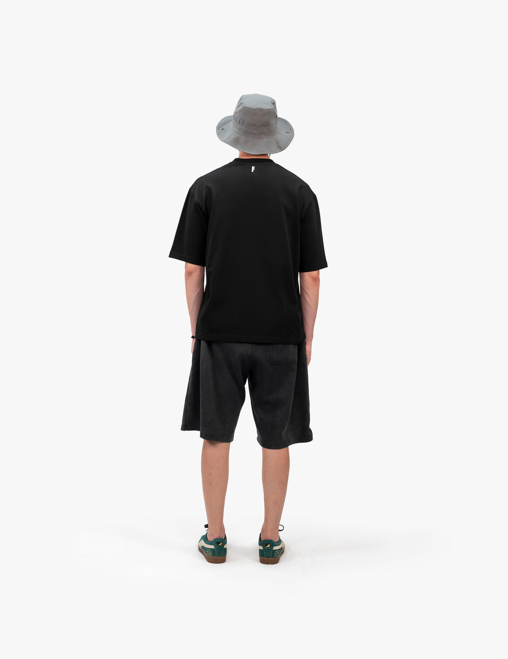 Team Summer 25 Siyah Oversize Tişört - Beyaz stüdyo fonunda, arkası dönük olarak poz veren erkek manken. Başında gri bir balıkçı şapkası (bucket hat) bulunmaktadır. Üzerindeki siyah oversize tişörtün ensesinin altında küçük, beyaz bir logo baskısı yer alıyor. Eskitme görünümlü antrasit rengi şort ve yeşil detaylı spor ayakkabılar kombini tamamlıyor. -  Faylit