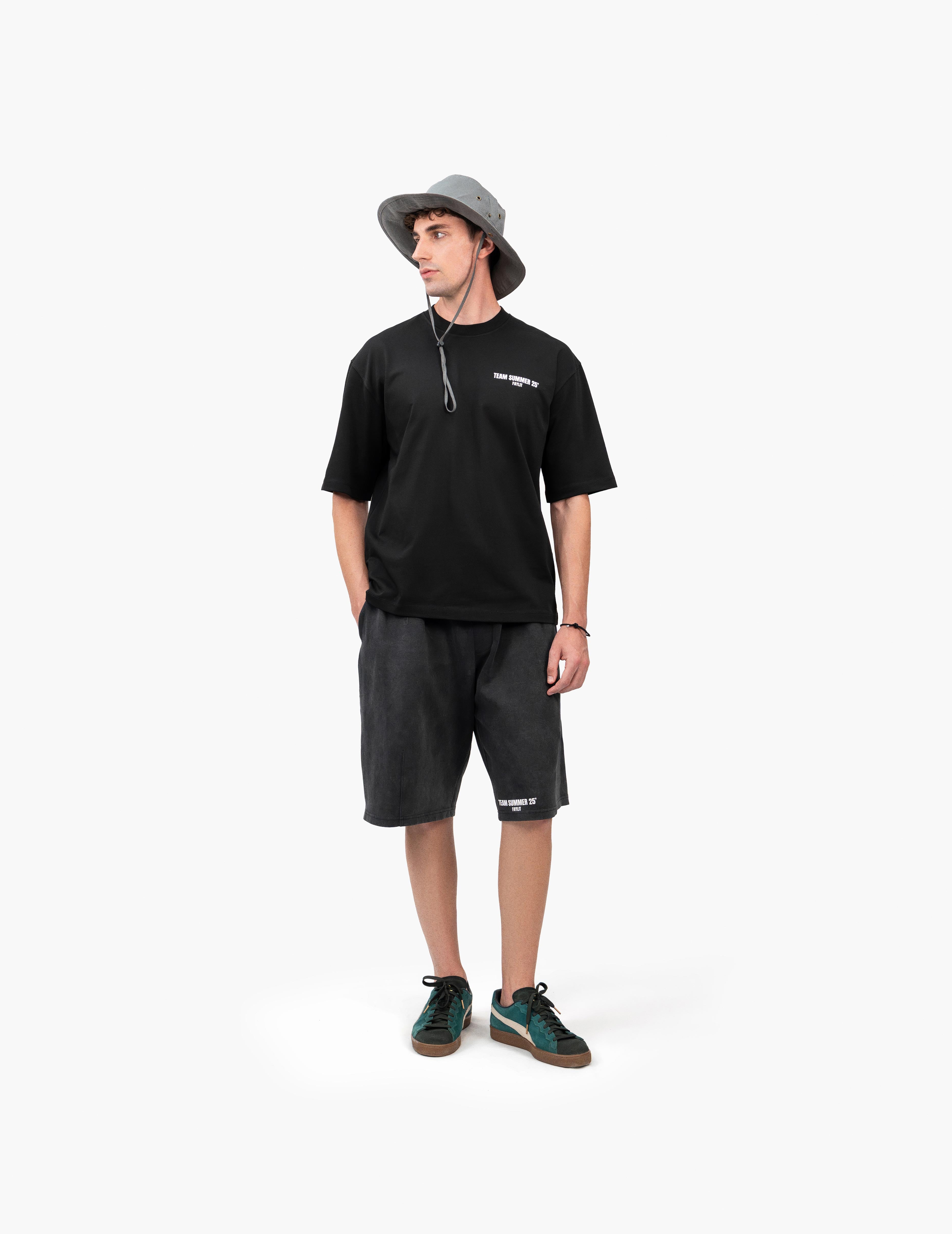 Team Summer 25 Siyah Oversize Tişört - Beyaz stüdyo fonunda, yana dönük poz veren erkek manken, outdoor ve sokak stilini yansıtan bir kombin sergiliyor. Başında ipli gri bir balıkçı şapkası, göğsünde beyaz yazı baskılı siyah oversize tişört ve eskitme görünümlü antrasit rengi şort giyiyor. Kombin, yeşil detaylı spor ayakkabılar ile tamamlanmıştır. Mankenin sağ eli şortunun cebindedir. -  Faylit