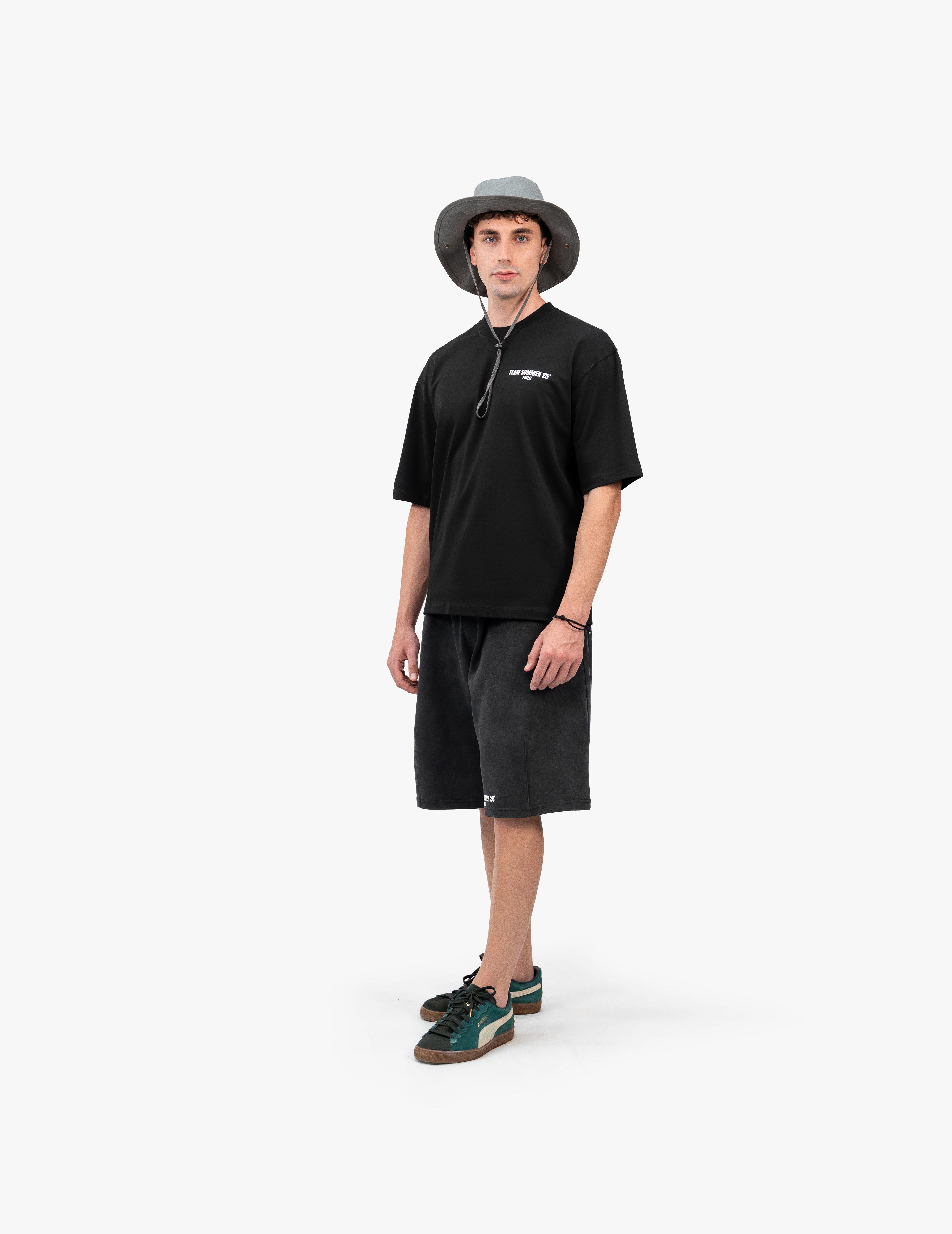 Team Summer 25 Siyah Oversize Tişört - Beyaz stüdyo fonunda, hafifçe yana dönmüş bir açıyla kameraya bakan erkek manken. Başında ipli gri bir balıkçı şapkası, üzerinde göğsünde ve şortunun paçasında beyaz yazı baskıları bulunan siyah oversize tişört ve antrasit rengi şort takımı giyiyor. Kombini, yanları yeşil detaylı, kahverengi tabanlı spor ayakkabılarla tamamlanmıştır. -  Faylit