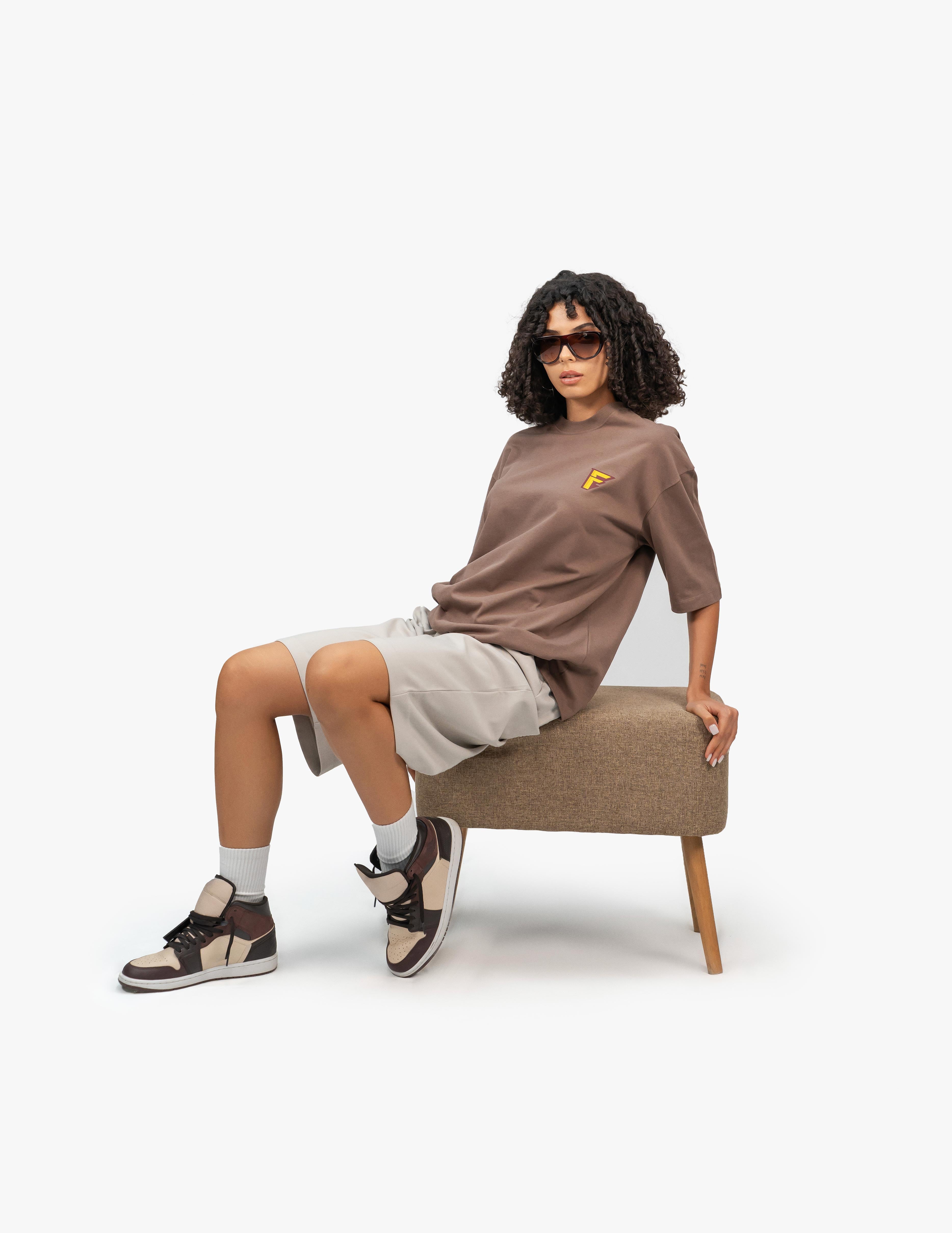 Thunder Kahverengi Oversize T-Shirt,  Beyaz bir stüdyo fonunun önünde, kahverengi bir taburede oturan, kısa siyah kıvırcık saçlı bir kadın. Siyah çerçeveli güneş gözlüğü takıyor ve kameraya bakıyor. Üzerinde göğüs kısmında sarı bir logo bulunan kahverengi, geniş kesim bir tişört ve bej renkli, diz hizasında bol bir şort giyiyor. Sol eli şortunun cebinde, sağ eli ise oturduğu taburenin kenarında duruyor. Ayaklarında bilekli spor ayakkabılar ve beyaz çoraplar bulunuyor. -  Faylit