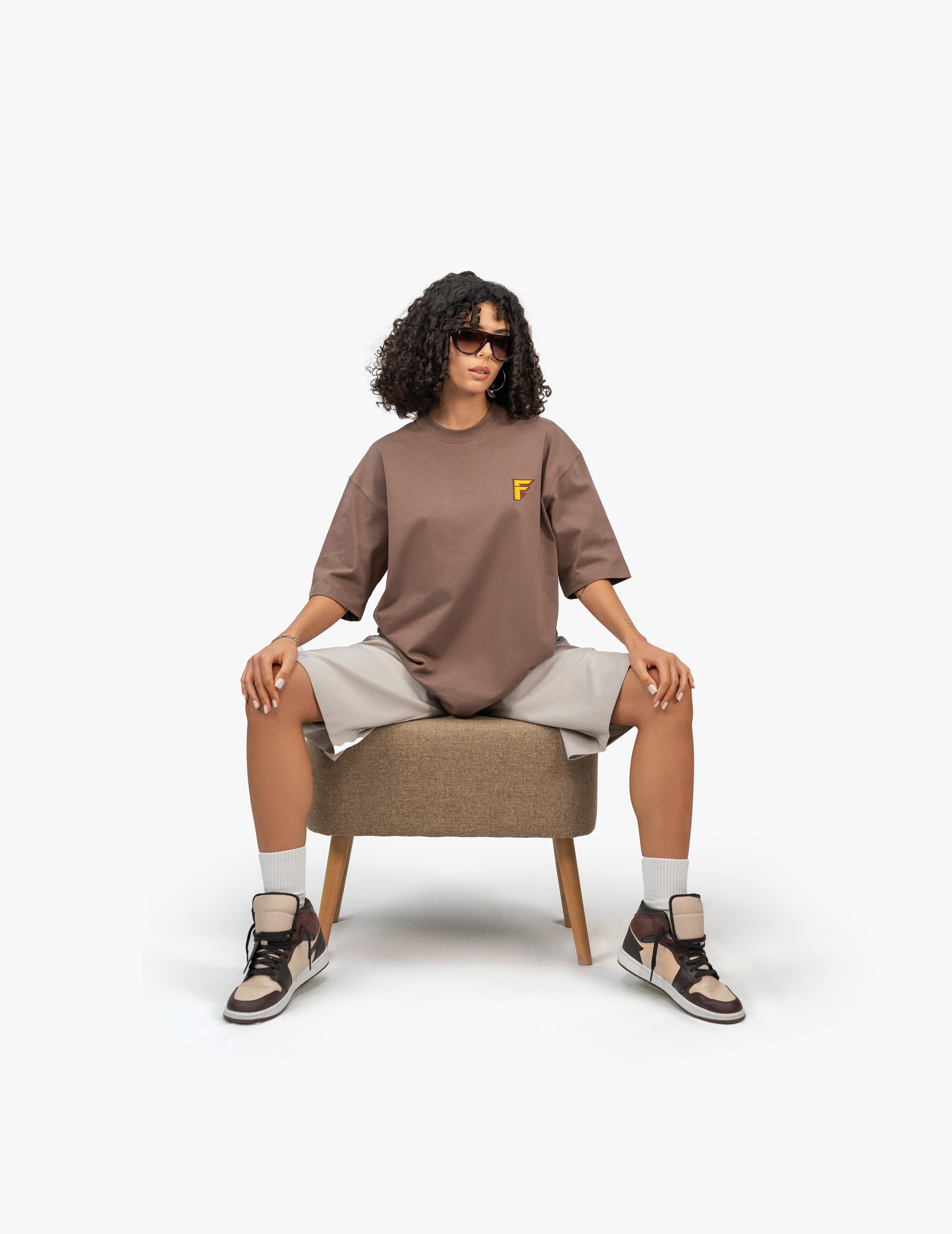 Thunder Kahverengi Oversize T-Shirt,  Beyaz bir stüdyo fonunun önünde, kahverengi bir taburede oturan, kısa siyah kıvırcık saçlı bir kadın. Siyah çerçeveli güneş gözlüğü takıyor ve bacakları omuz genişliğinde açık. Üzerinde göğüs kısmında sarı bir logo bulunan kahverengi, geniş kesim bir tişört ve bej renkli, diz hizasında bol bir şort giyiyor. Ayaklarında bilekli spor ayakkabılar ve beyaz çoraplar bulunuyor. -  Faylit