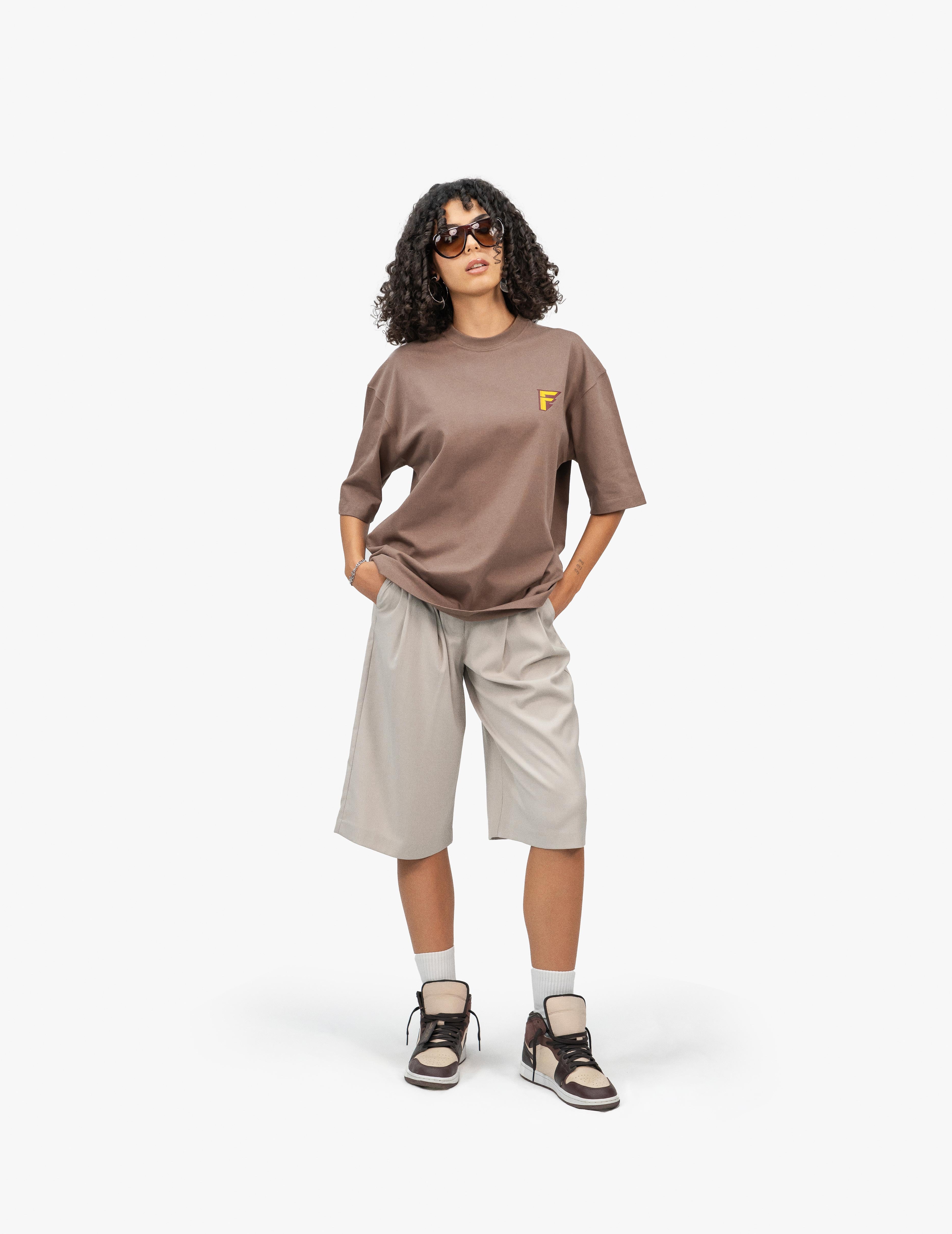 Thunder Kahverengi Oversize T-Shirt, Beyaz bir stüdyo fonunun önünde, hafifçe sağa dönük duran, kısa siyah kıvırcık saçlı bir kadın. Siyah çerçeveli güneş gözlüğü takıyor, elleri ceplerinde ve doğrudan kameraya bakıyor. Üzerinde göğüs kısmında sarı bir logo bulunan kahverengi, geniş kesim bir tişört ve bej renkli, diz hizasında bol bir şort giyiyor. Ayaklarında bilekli spor ayakkabılar ve beyaz çoraplar bulunuyor. -  Faylit