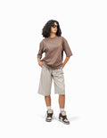 Thunder Kahverengi Oversize T-Shirt,  Beyaz bir stüdyo fonunun önünde, hafifçe sağa dönük duran, kısa siyah kıvırcık saçlı bir kadın. Siyah çerçeveli güneş gözlüğü takıyor ve elleri ceplerinde. Üzerinde göğüs kısmında sarı bir logo bulunan kahverengi, geniş kesim bir tişört ve bej renkli, diz hizasında bol bir şort giyiyor. Ayaklarında bilekli spor ayakkabılar ve beyaz çoraplar bulunuyor. -  Faylit